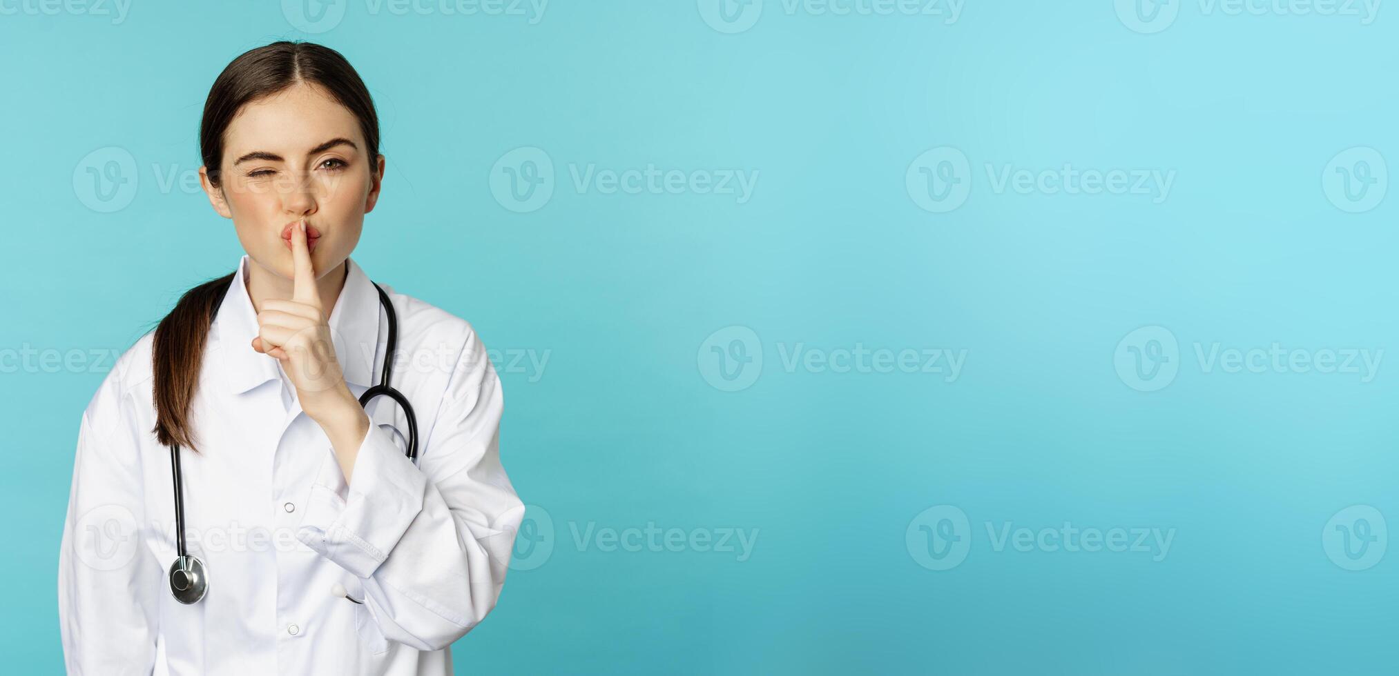 Smiling cute doctor woman, shush, press hush shh sign with finger pressed to lips, hiding secret, taboo gesture, standing over torquoise background photo