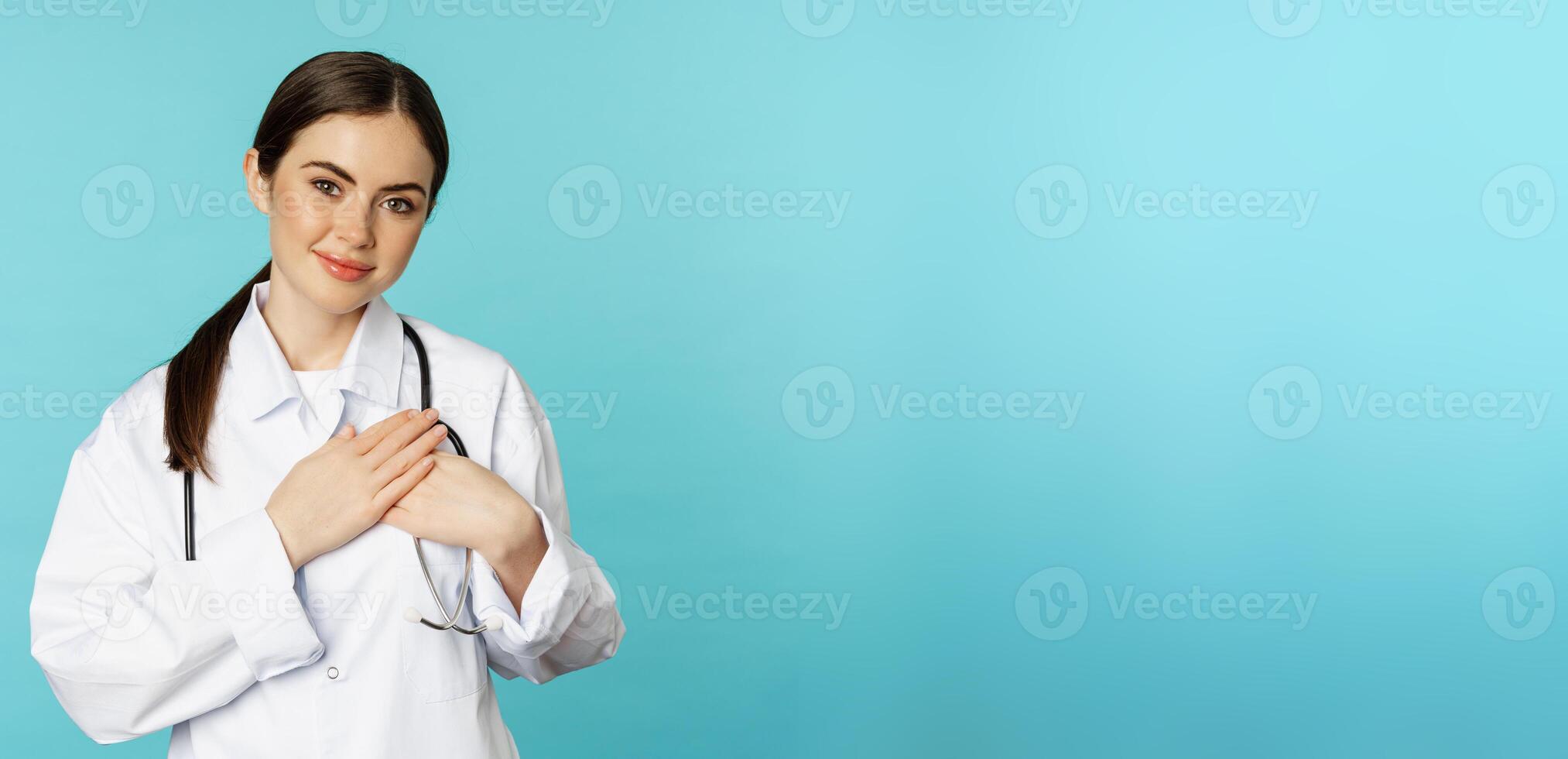 Caring healthcare worker, young female doctor holding hands on heart and smiling, looking with care and tenderness, standing over torquoise background photo