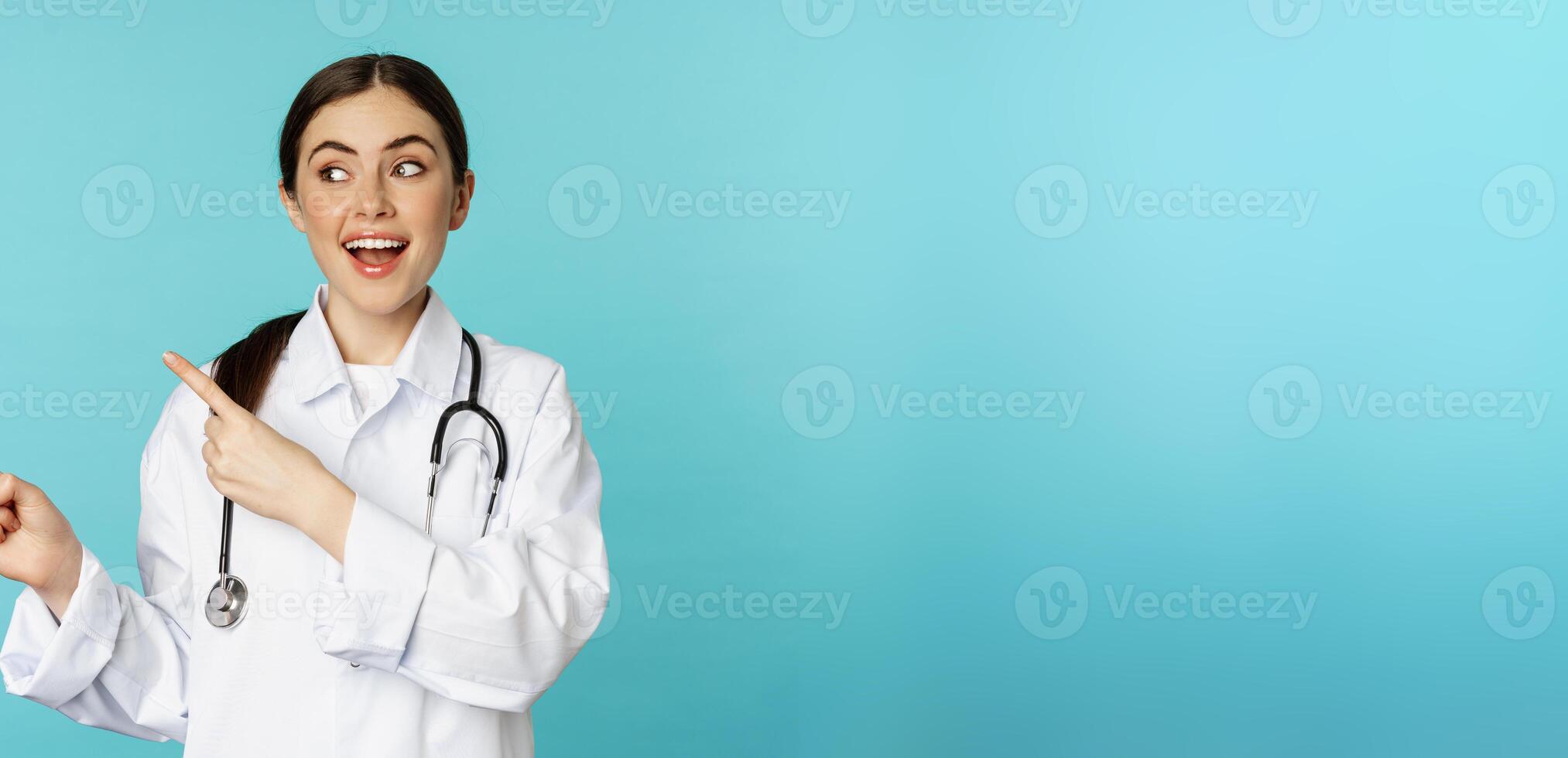 Portrait of smiling medical worker, girl doctor in white coat with stethoscope, pointing fingers left, showing medical clinic advertisement, torquoise background photo