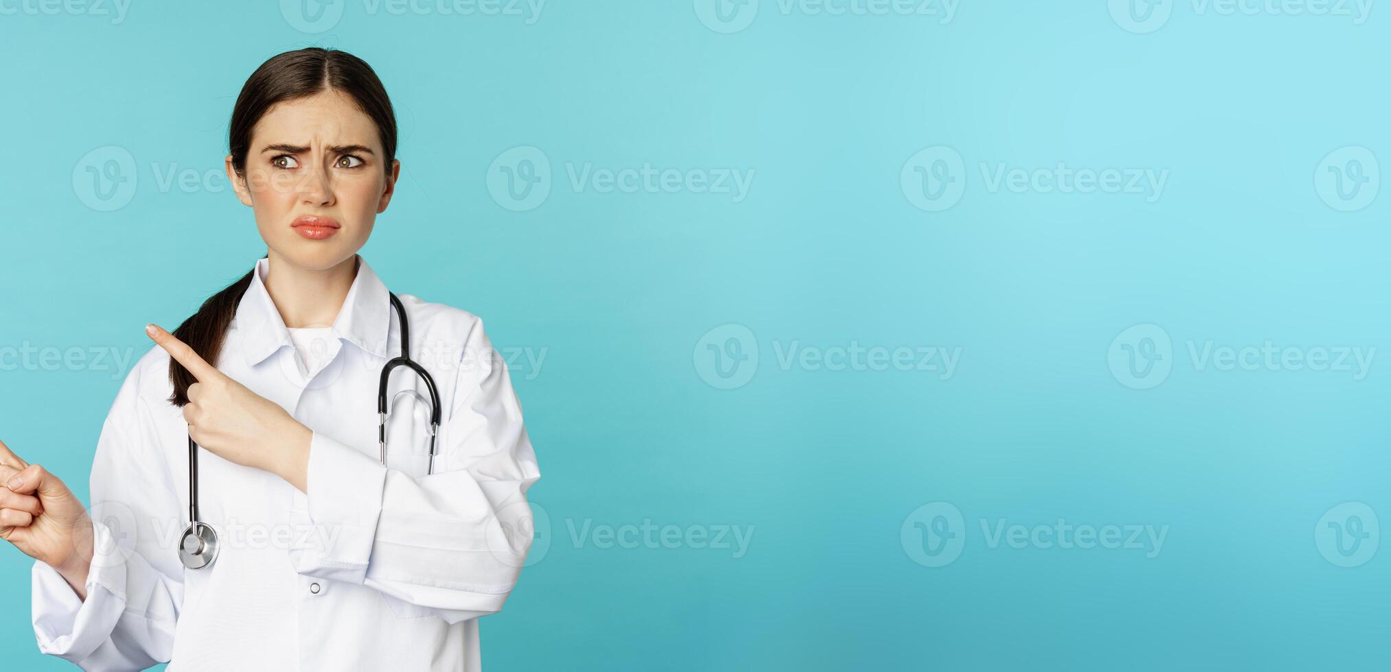 Confused, disappointed medical worker, frowning while pointing fingers left with doubtful, puzzled face expression, standing over torquoise background photo