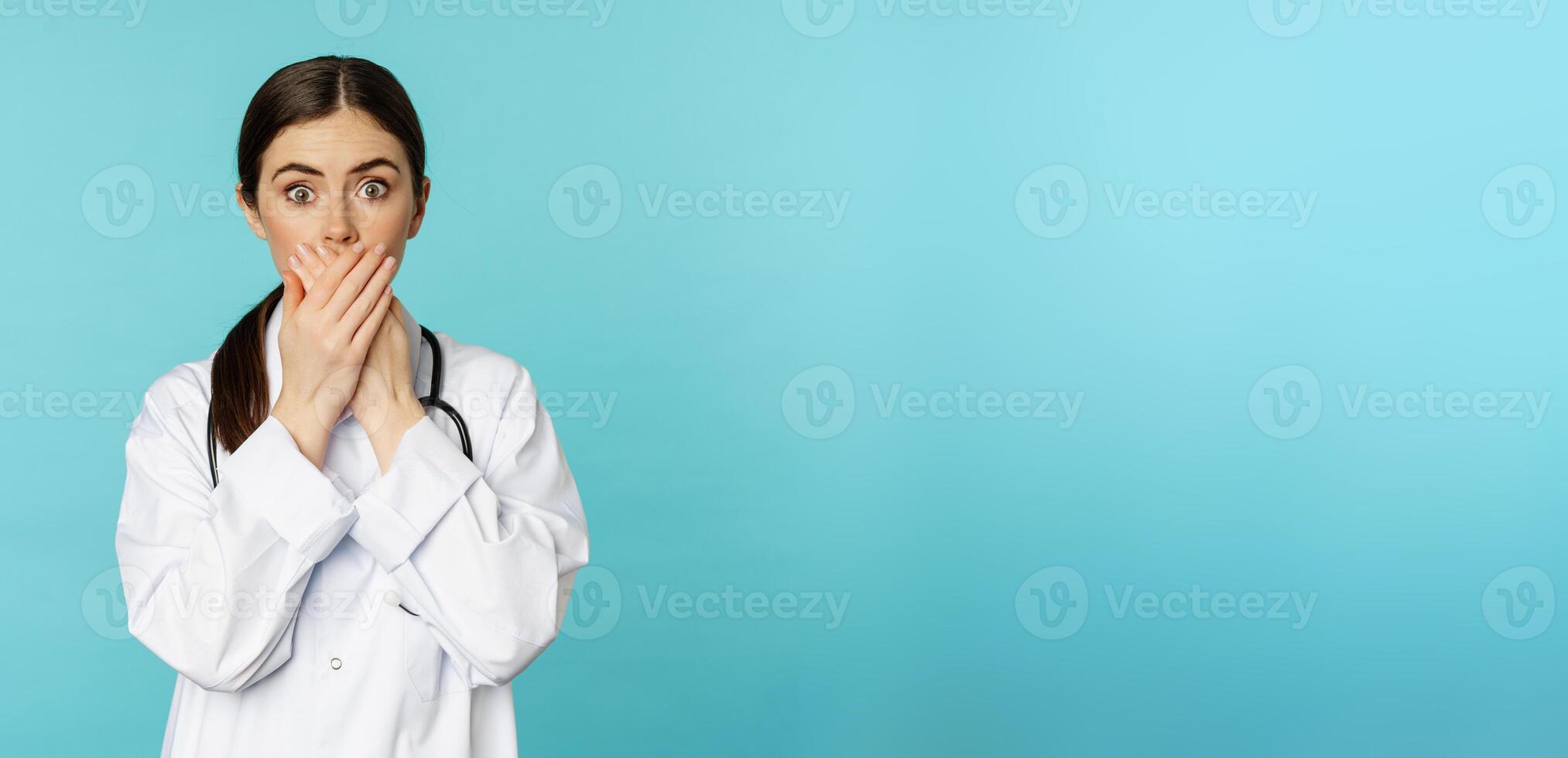 Shocked and concerned woman doctor, clinic worker, covering mouth with hands, looking startled at camera, standing over blue background photo