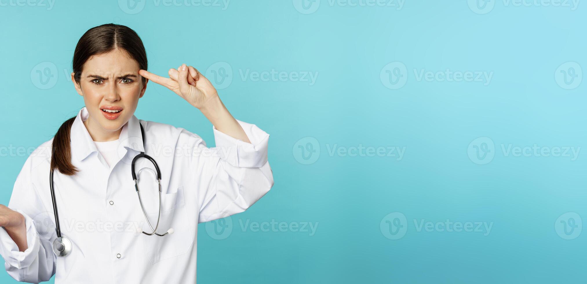 Annoyed woman doctor pointing finger at head, scolding someone stupid, crazy or strange, standing over torquoise background photo