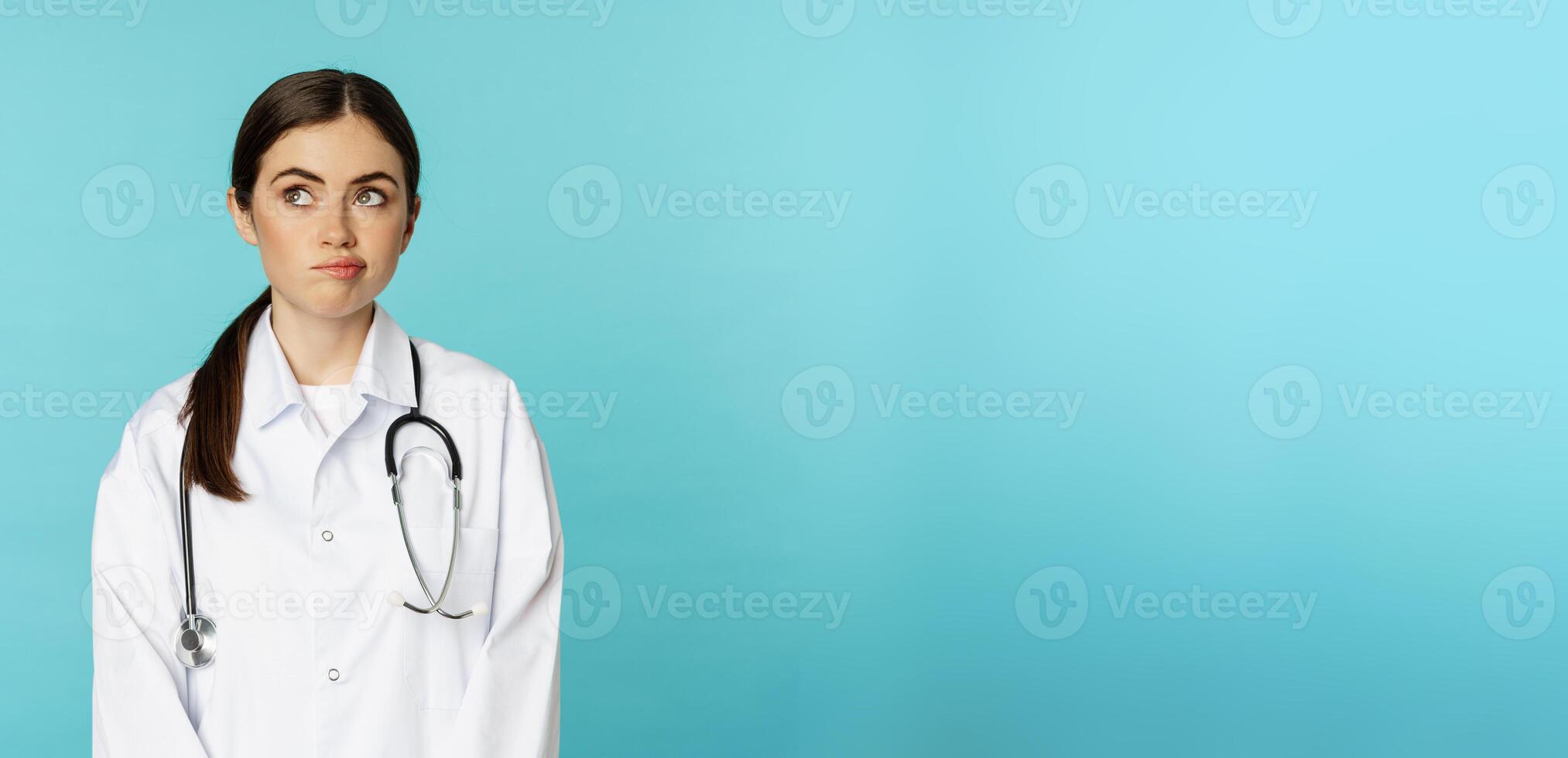 Image of woman doctor, female medial staff in white lab coat, looking away thoughful, making decision, thinking of smth, standing over blue background photo