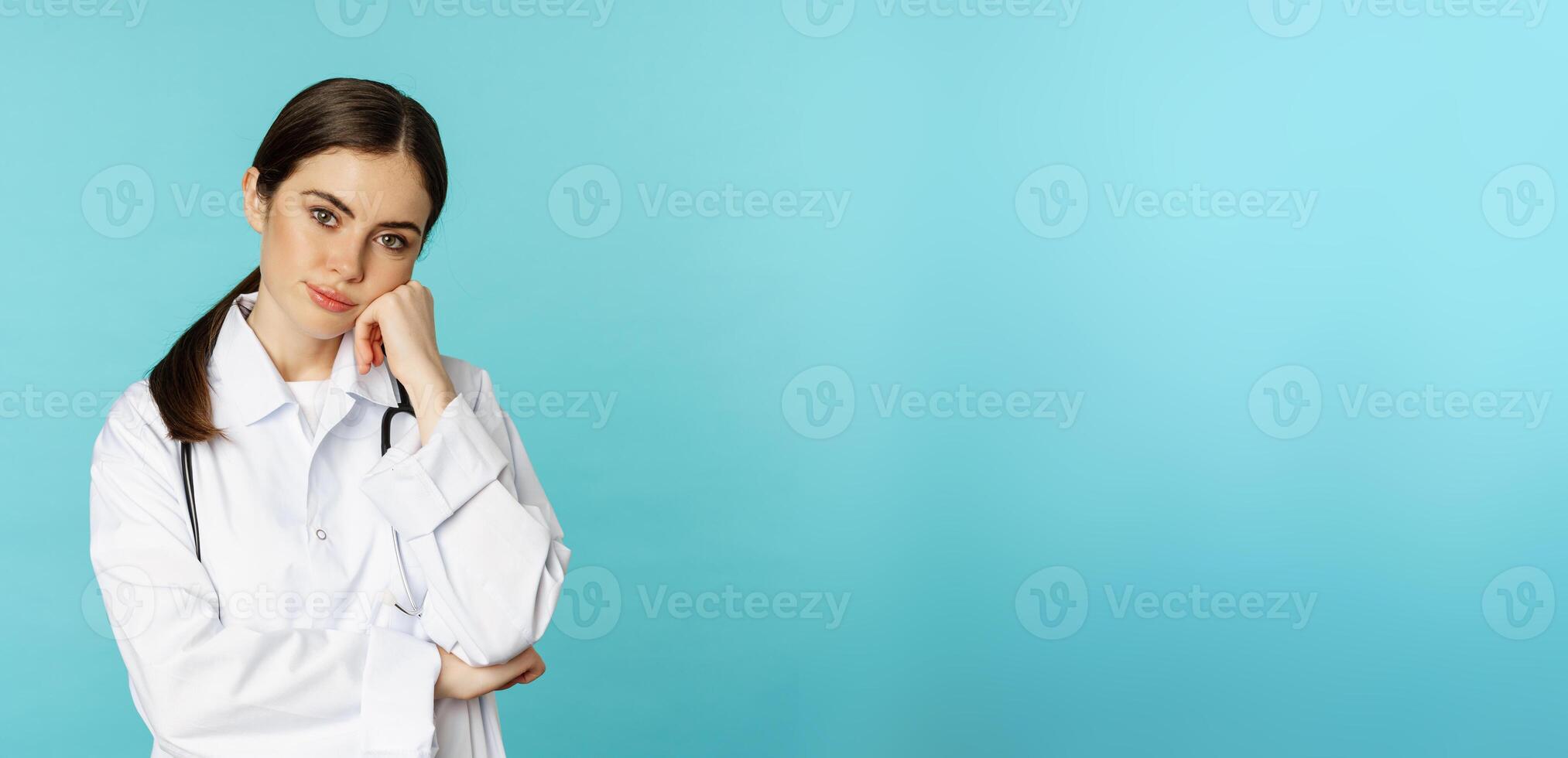Thoughtful medical worker, young doctor woman looking unamused, bored, standing in white coat against blue background photo