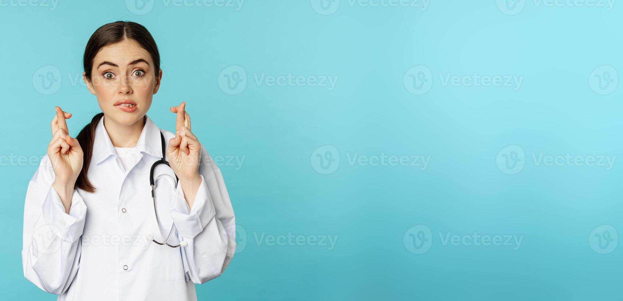 Hopeful woman doctor, lab worker cross fingers and smiling, praying, making wish, anticipating, standing over torquoise background photo