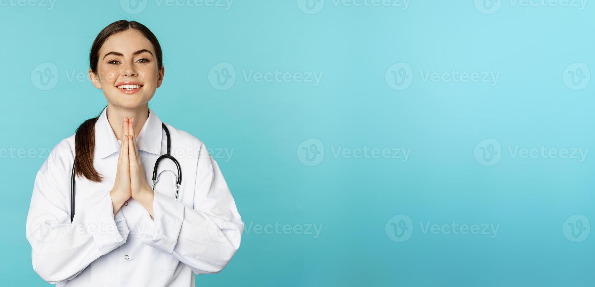 Smiling hopeful young woman doctor, holding hands in begging, thank you pose, standing over blue background photo