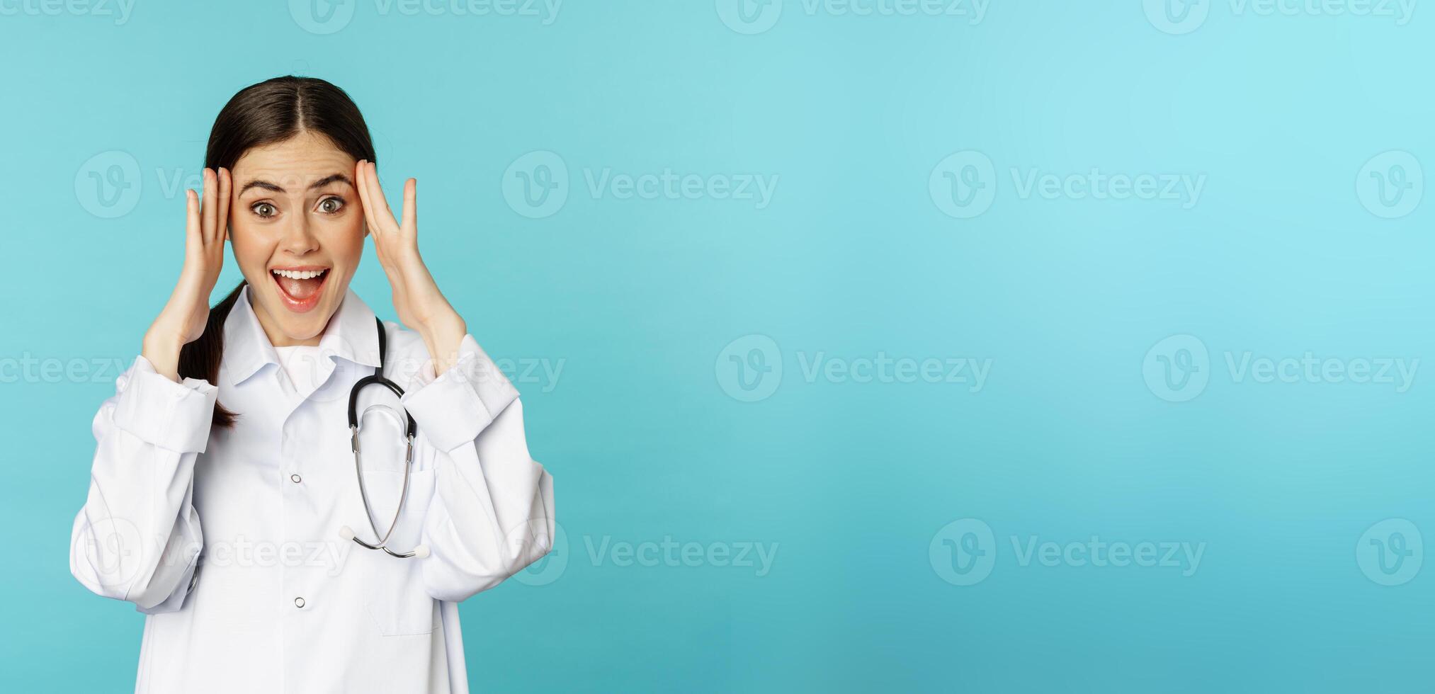 Portrait of surprised and excited smiling doctor woman, holding hands on head, reaction to mindblowing awesome news, standing over blue background photo