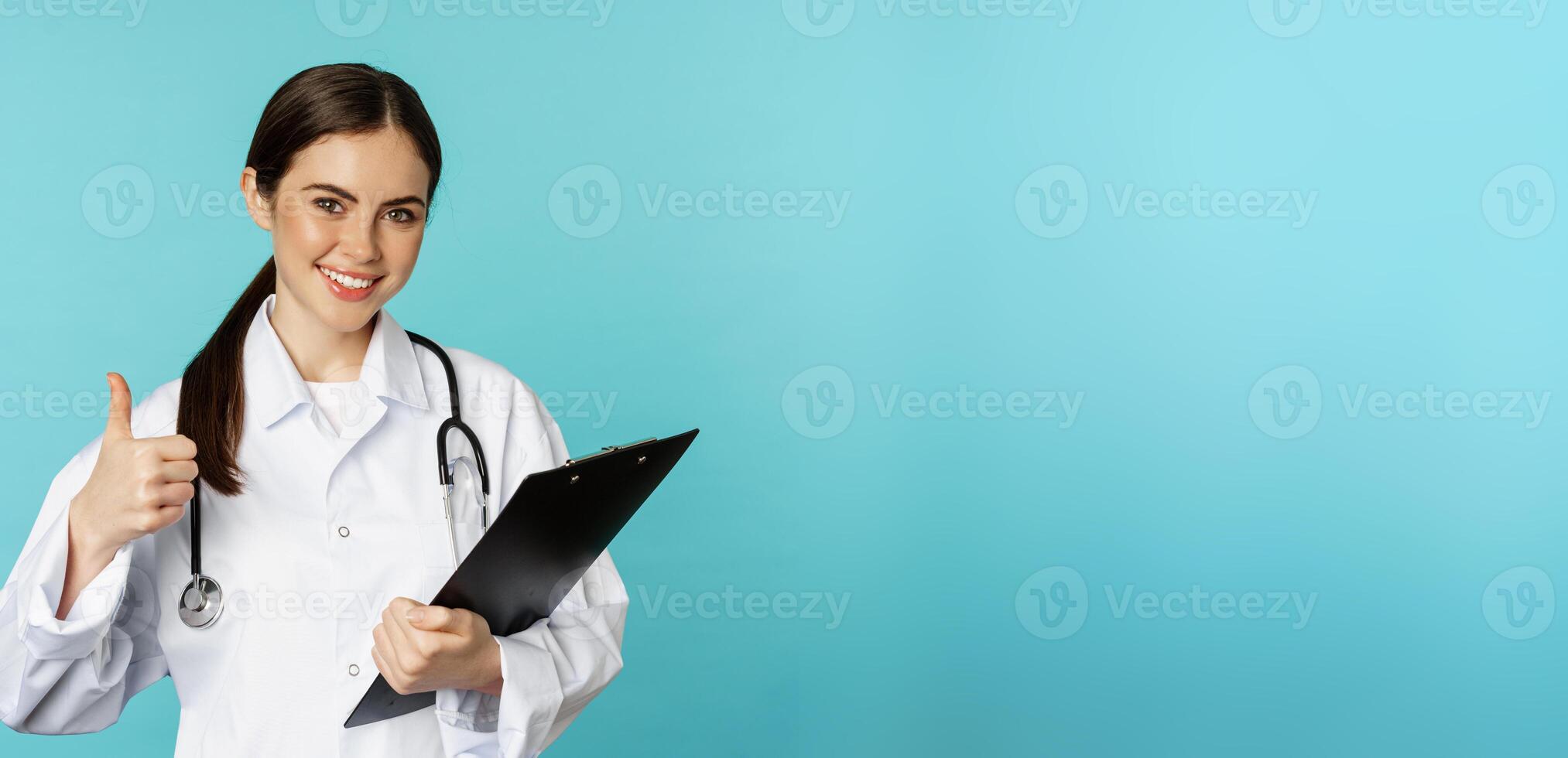 Confident smiling doctor woman, physician showing thumbs up, holding clipboard, appointment in hospital, standing over torquoise background photo
