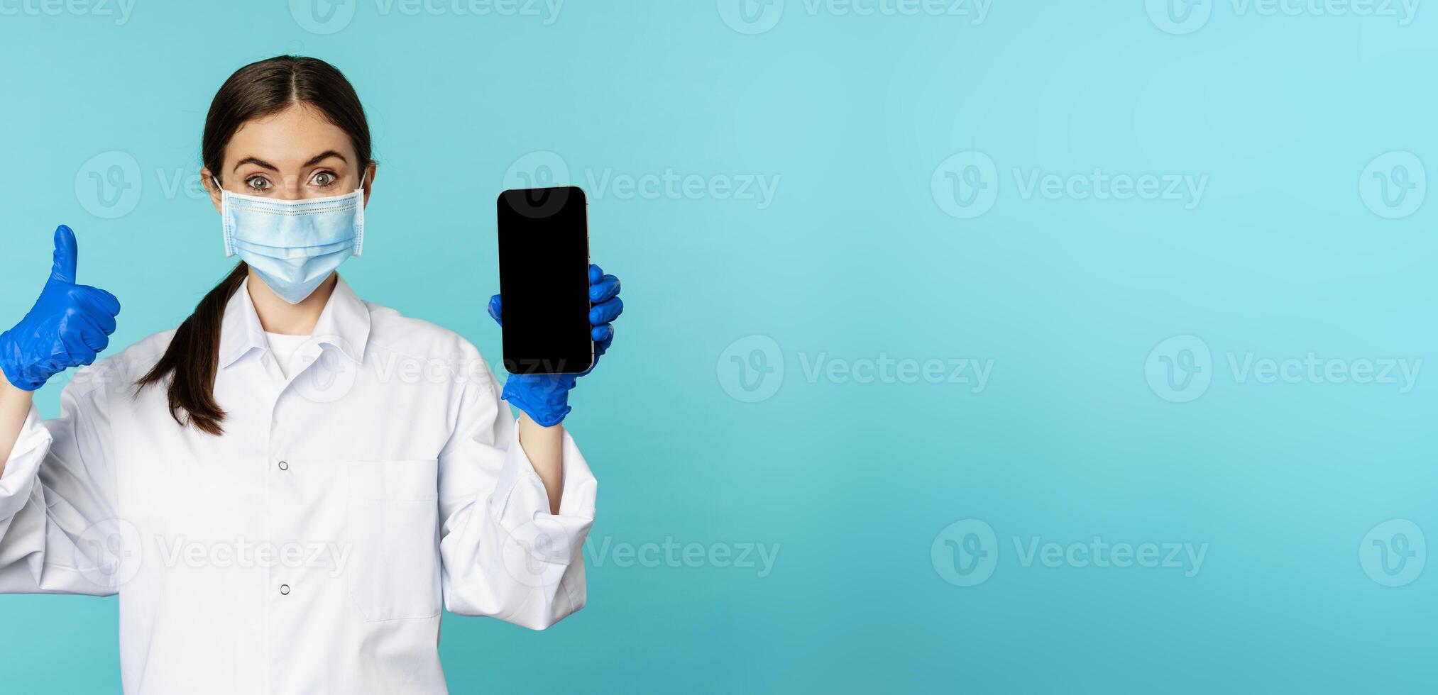 Portrait of doctor in medical face mask and gloves, showing mobile phone app, smartphone screen and thumb up, recommending online checkup website, standing over blue background photo