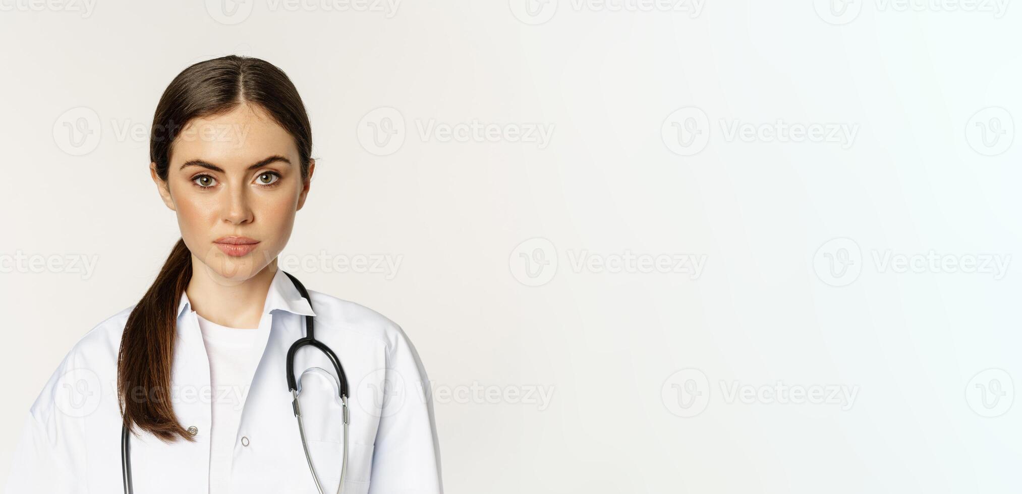Close up portrait, face of young woman doctor with stethoscope, looking serious, professional healthcare worker in clinic, standing over white background photo