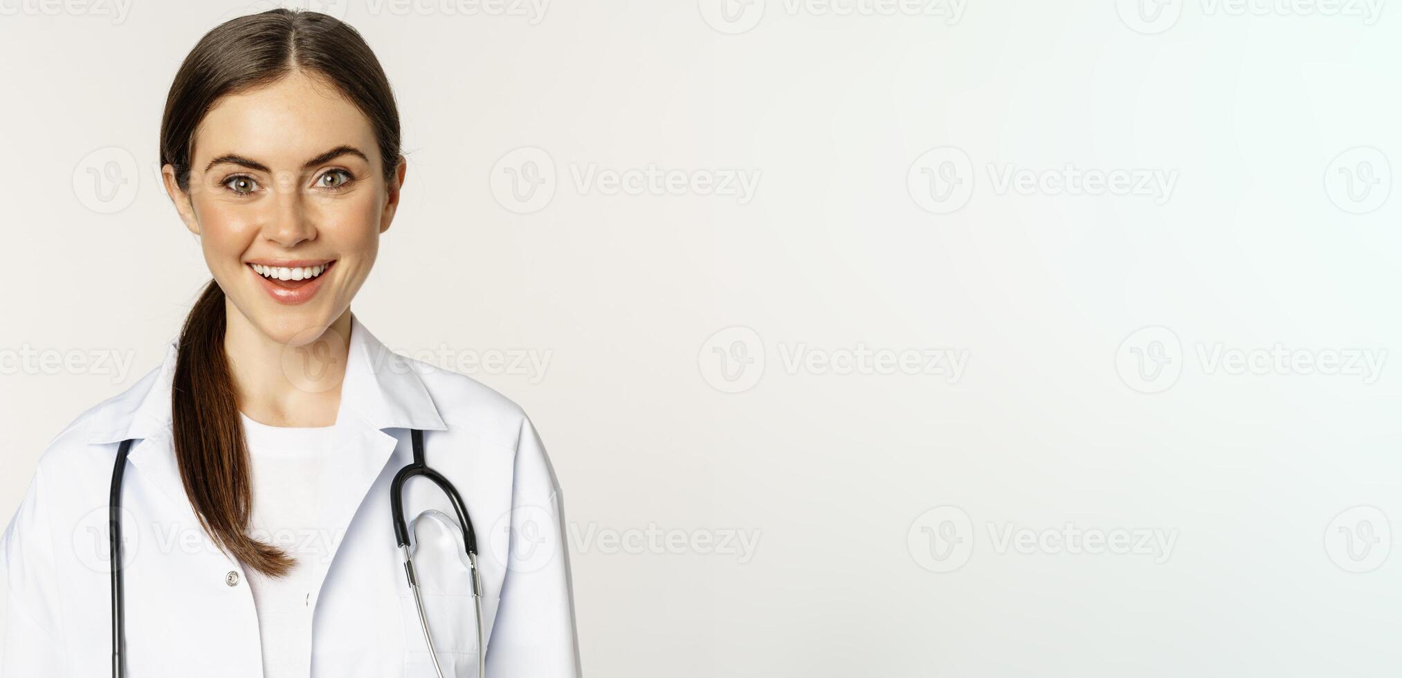 Smiling woman doctor, physician at appointment, looking happy and confident, wearing white coat and stethoscope, standing against studio background photo