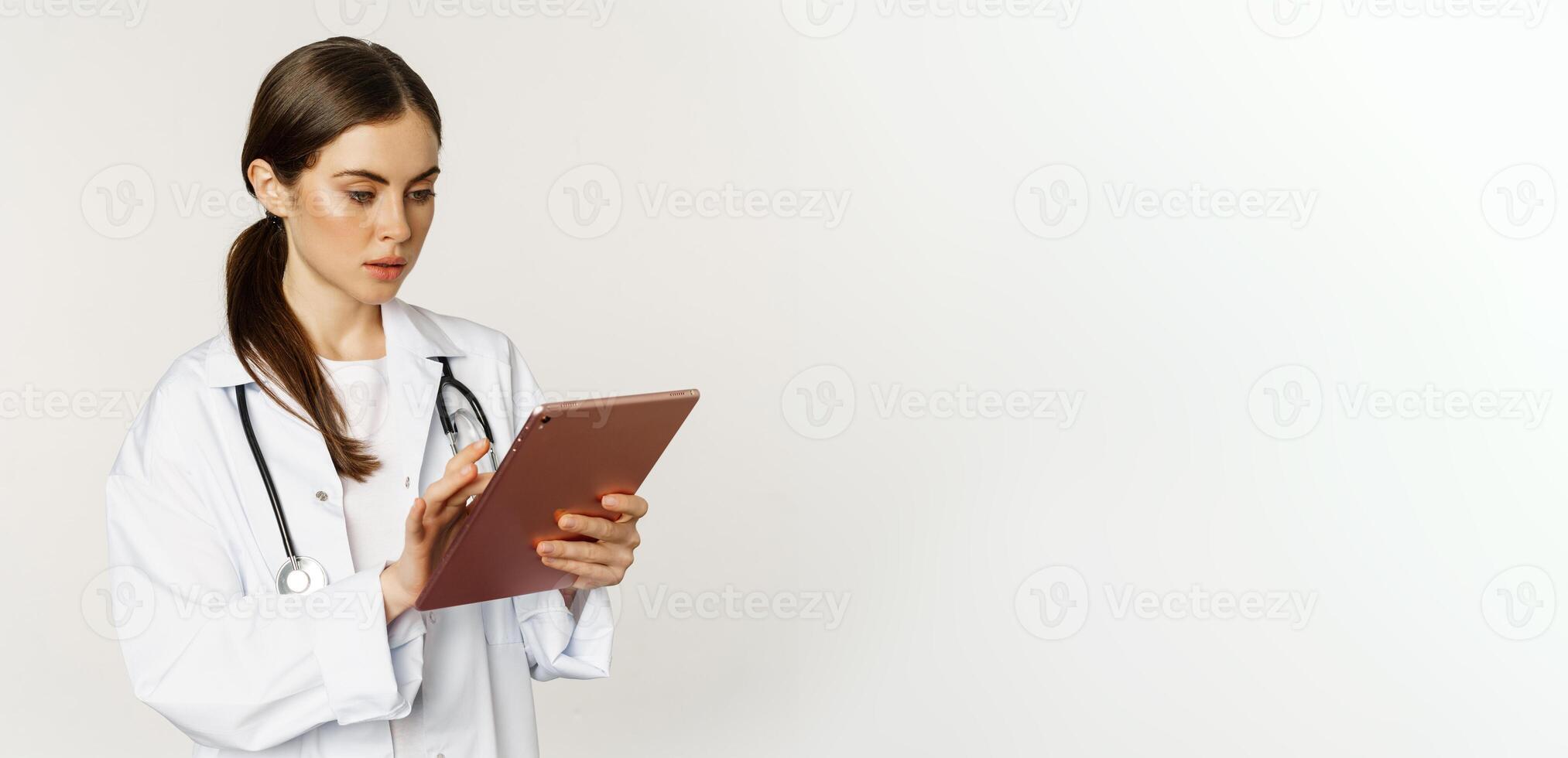Woman doctor looking concerned at digital tablet, reading with worried face expression, wearing white coat, standing over white background photo