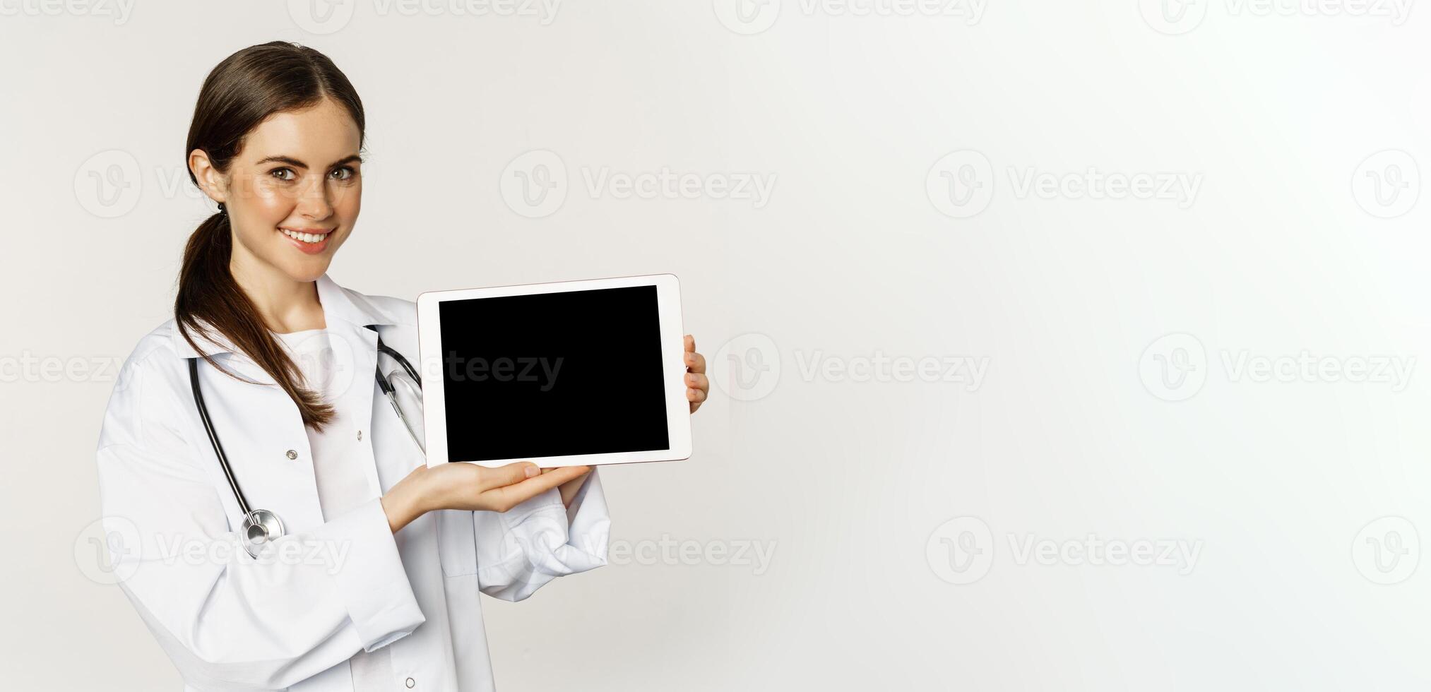Image of woman doctor, female healthcare worker showing online medical website, digital tablet screen and smiling, standing in white coat over white background photo