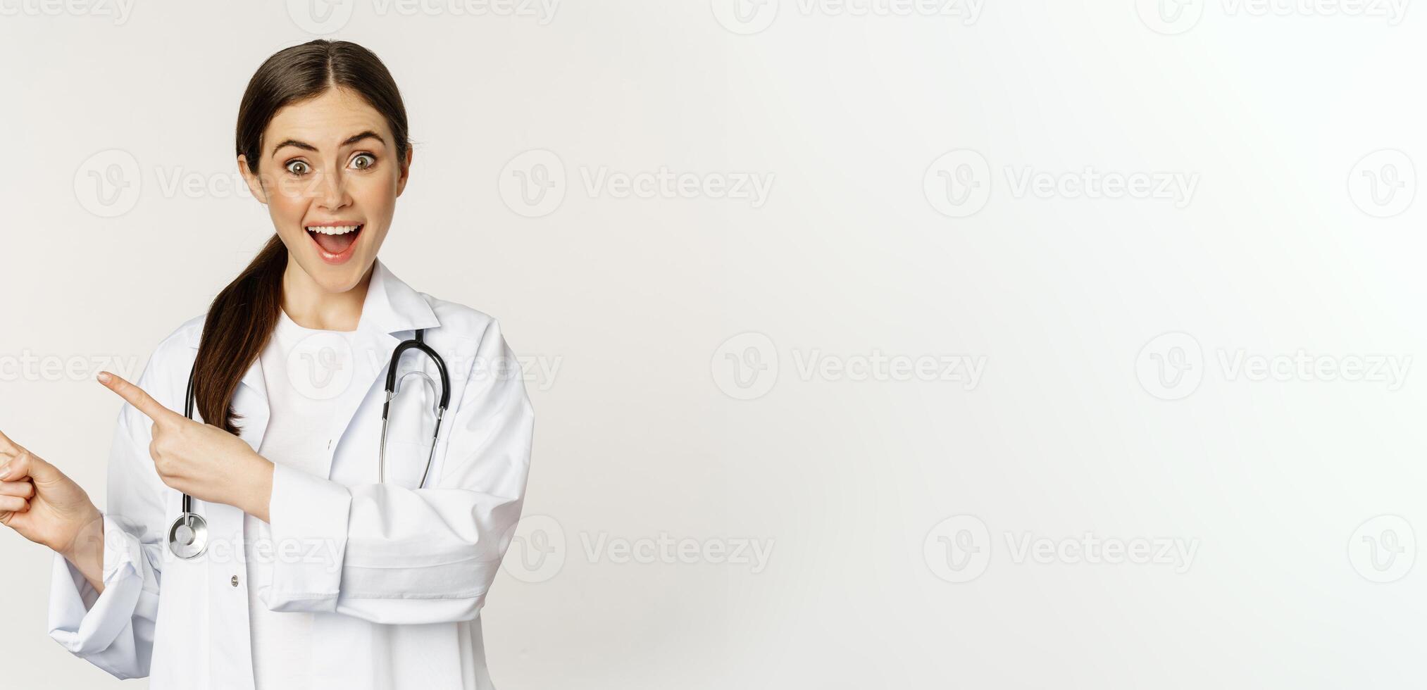 Portrait of smiling young woman doctor, healthcare medical worker, pointing fingers left, showing clinic promo, logo or banner, standing over white background photo