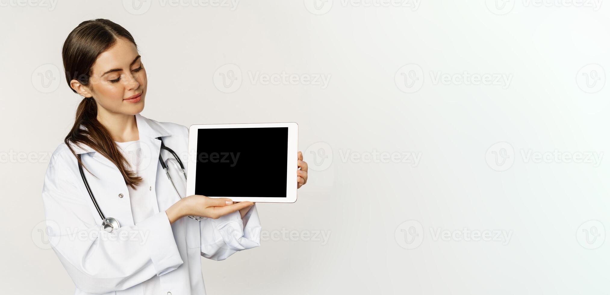 Image of woman doctor, female healthcare worker showing online medical website, digital tablet screen and smiling, standing in white coat over white background photo