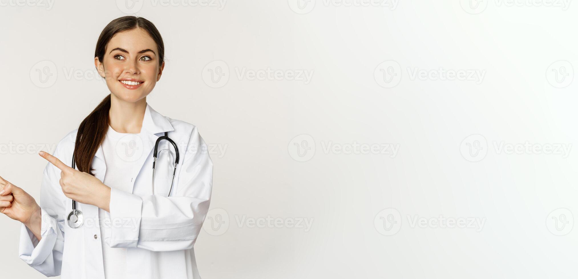 Portrait of smiling young woman doctor, healthcare medical worker, pointing fingers left, showing clinic promo, logo or banner, standing over white background photo