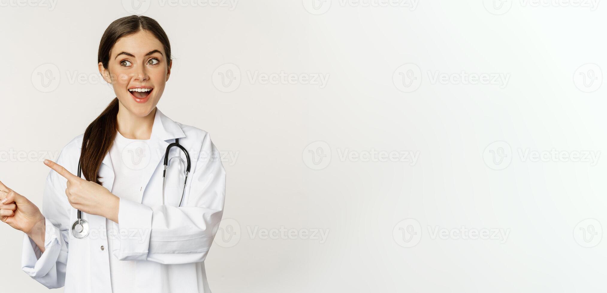 Surprised doctor, physician pointing fingers left and looking at logo banner, showing clinic advertisement, standing over white background photo