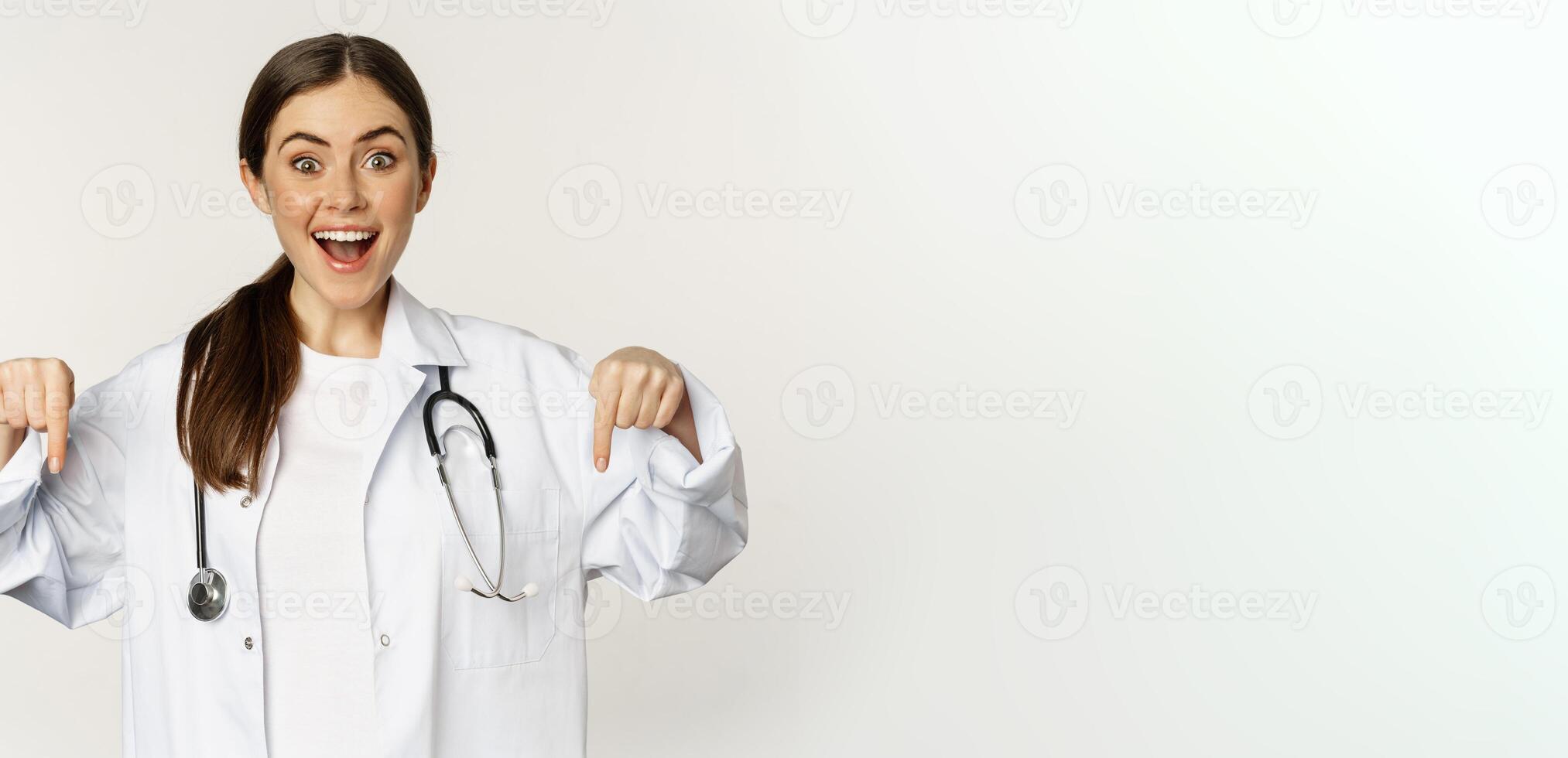 Portrait of happy female doctor, pointing fingers down and smiling, demonstrating promo offer, discount, standing over white background photo