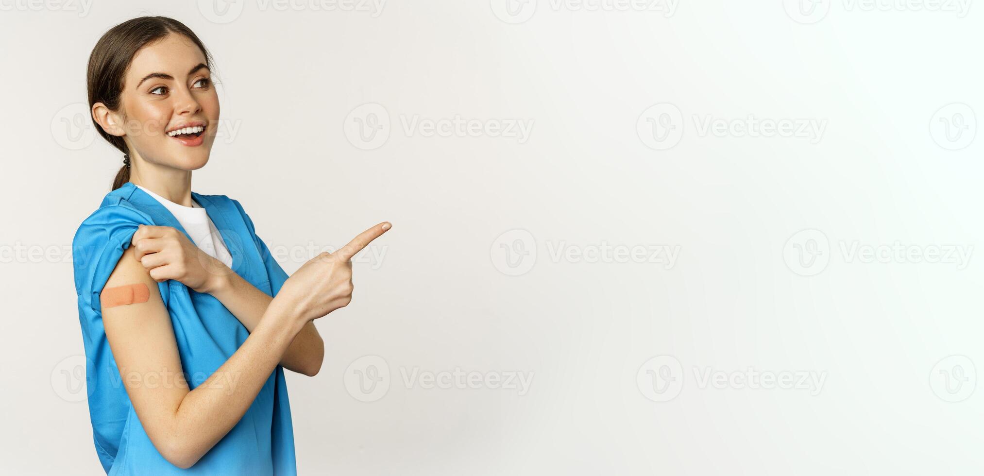 Smiling nurse, female healthcare worker in scrubs, pointing finger right, showing patch on shoulder, vaccination campaign, covid-19 and pandemic concept, white background photo