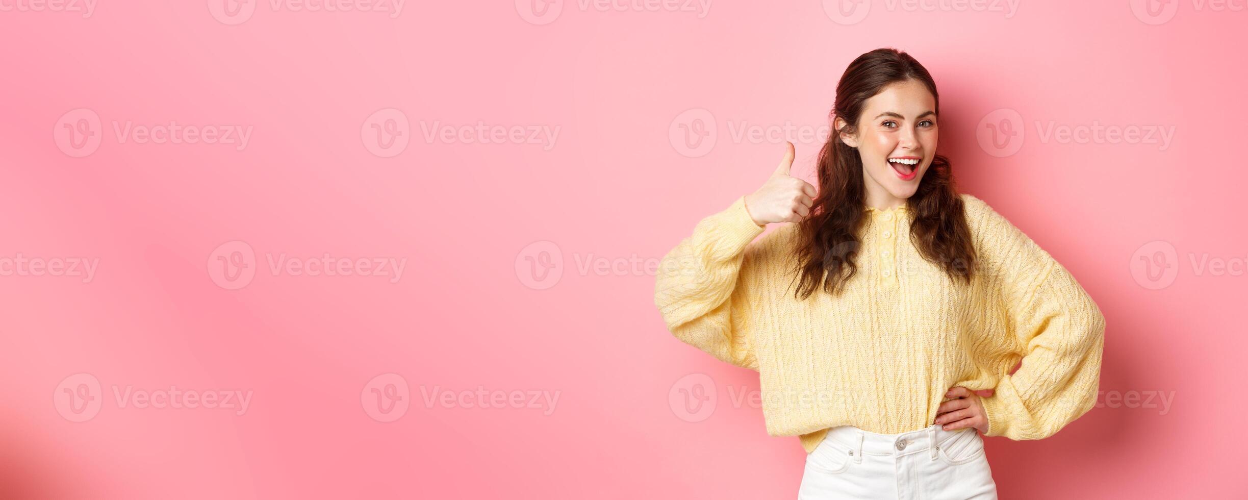 Portrait of beautiful brunette female model showing thumbs up, like your idea, give support, approve or praise something good, standing against pink background photo