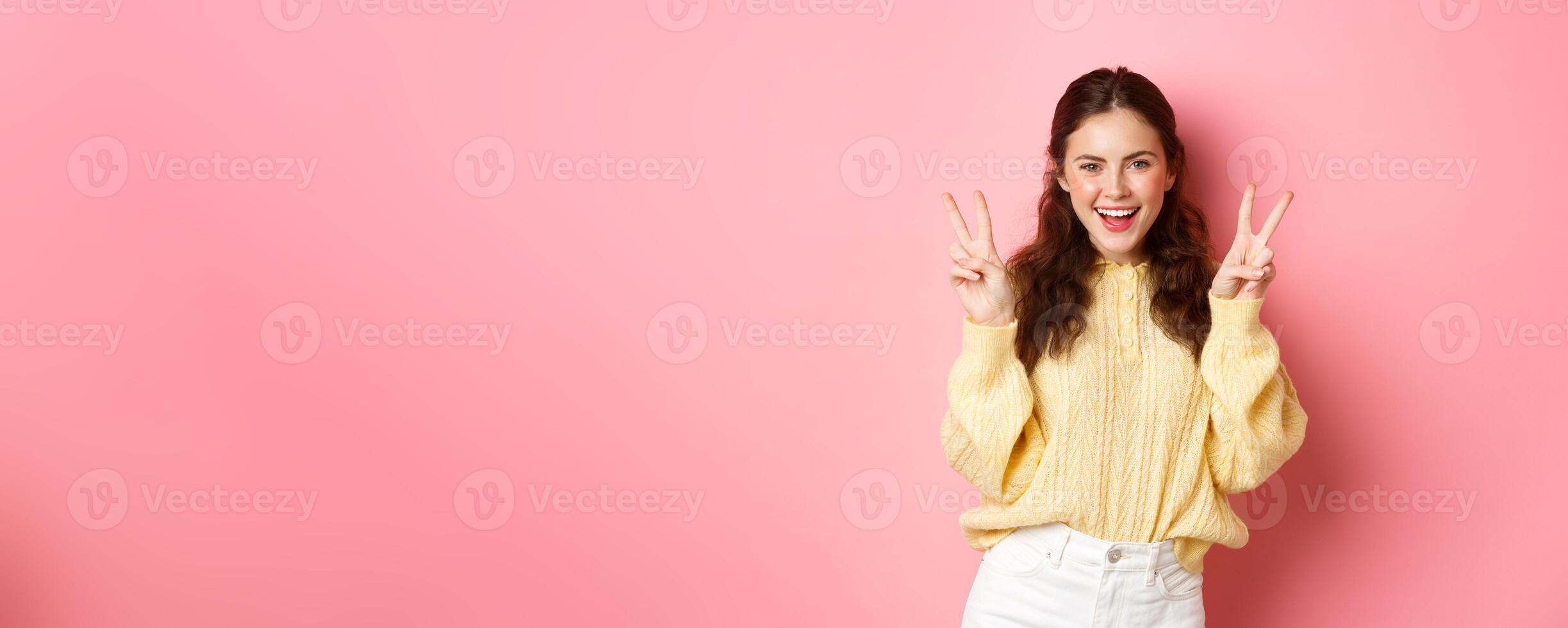 Portrait of attractive brunette female model, smiling and showing peace signs, making v-sign gesture, standing positive and happy against pink background photo