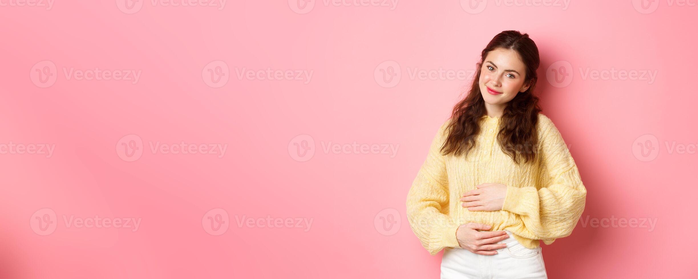 Young smiling woman being pregnant, rubbing her belly and looking with hopeful happy face at camera, expecting for baby, standing against pink background photo