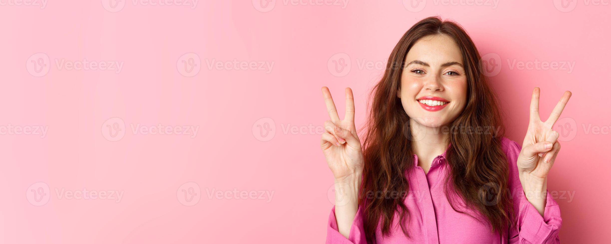 Close up of positive brunette female model showing peace signs, smiling and having fun, good vibes only, standing over pink background photo