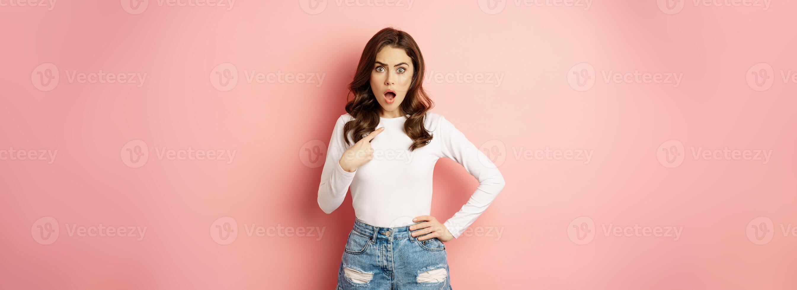 Shocked and offended young beautiful woman, pointing finger at herself, oh my gosh, who me gesture, being insulted, standing in disbelief over pink background photo