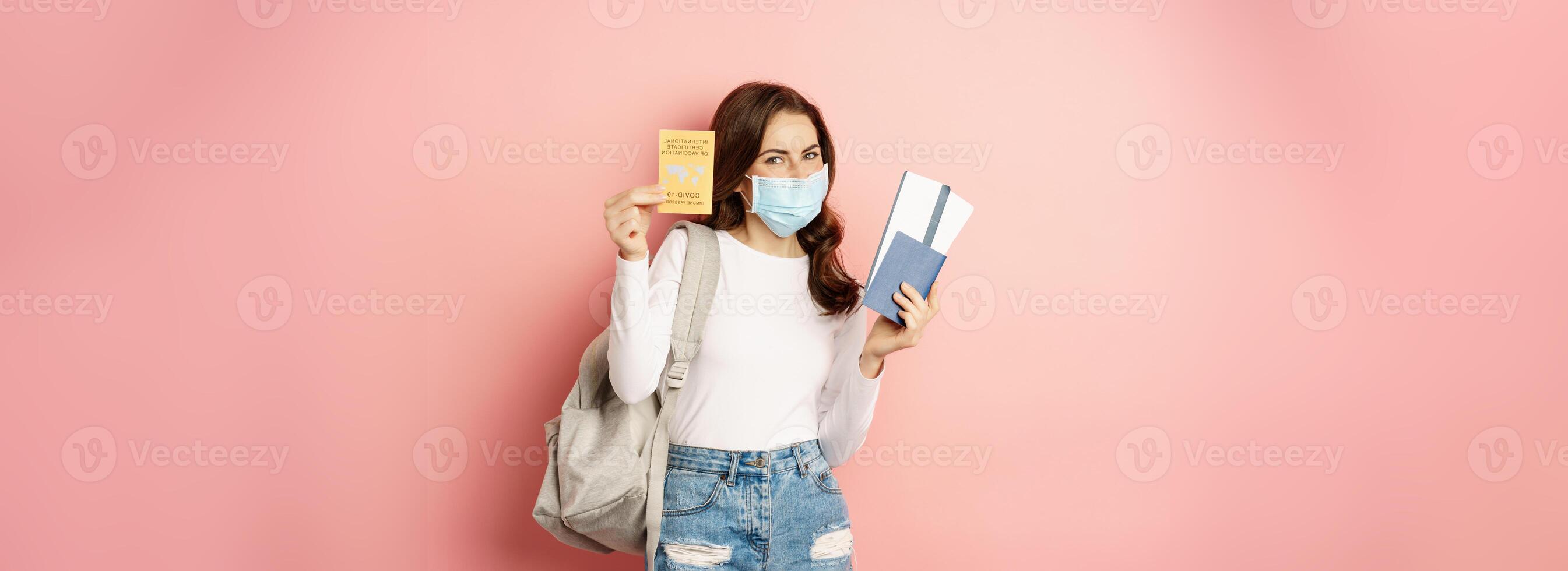Young woman going on trip, wearing medical mask, showing covid certificate, passport and aiplance tickets, vacation during pandemic, pink background photo