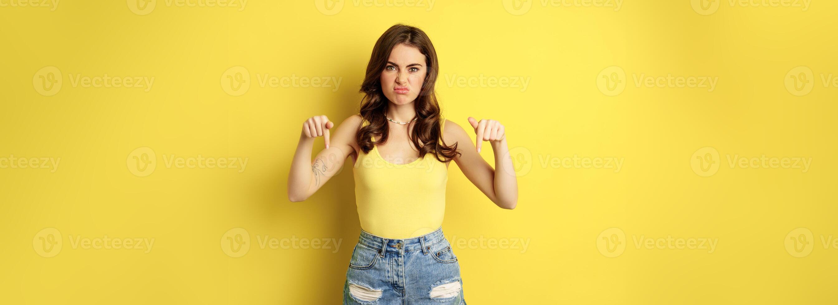 Disappointed girl grimacing, pointing fingers, showing logo, promo offer, banner or logo, standing over yellow background photo