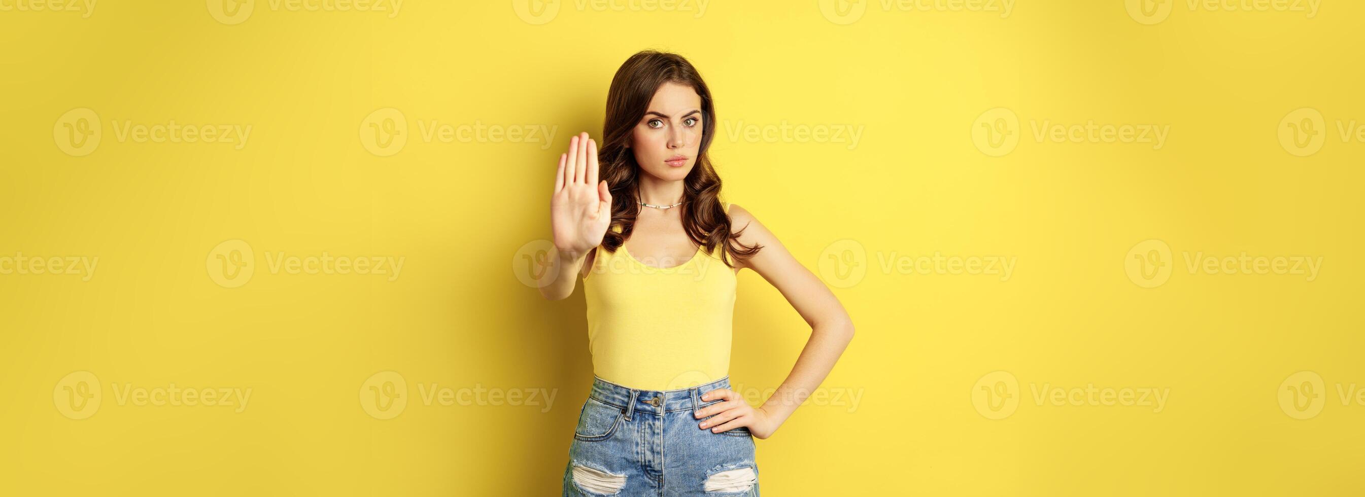 Serious stylish woman showing stop, rejection, prohibition gesture, decline something, disapprove, standing over yellow background photo