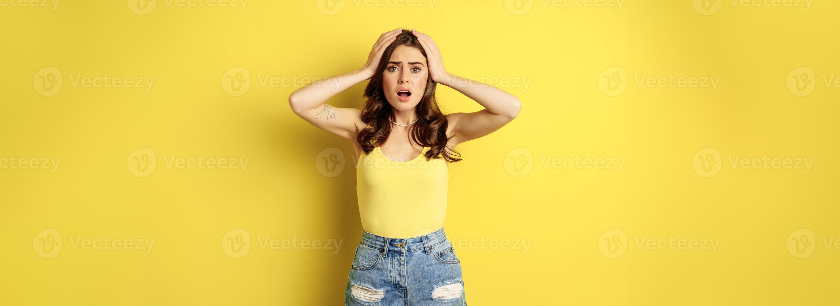 Troubled and shocked woman holding hands on head, panicking, feeling distressed and anxious, having problem, standing over yellow background photo