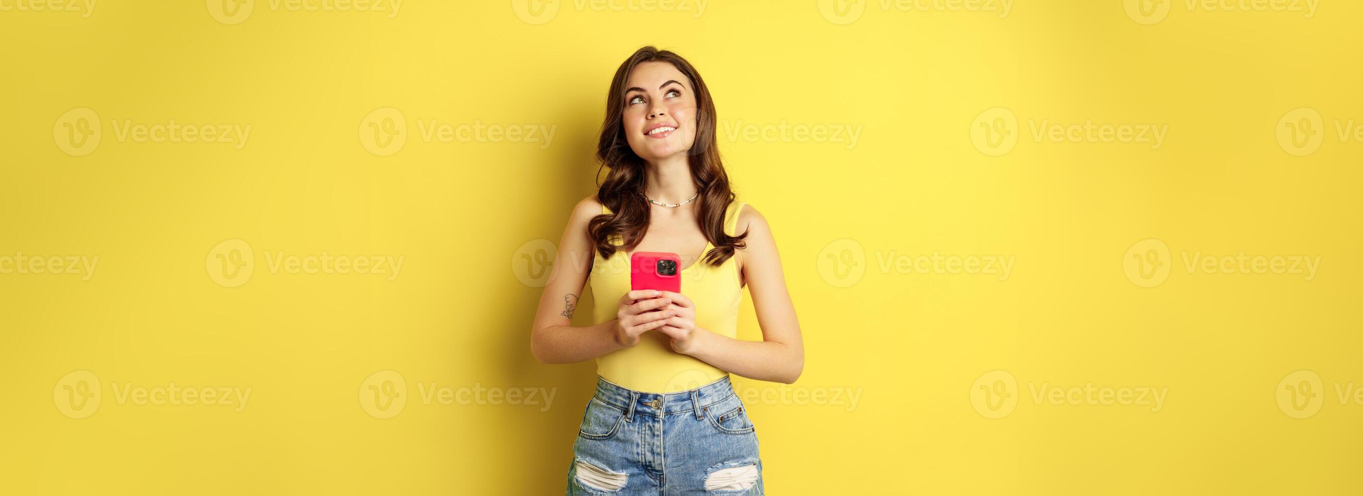 Technology and cellphone. Beautiful stylish girl order in online store, praying in application, using smartphone app and thinking, making decision, standing over yellow background photo