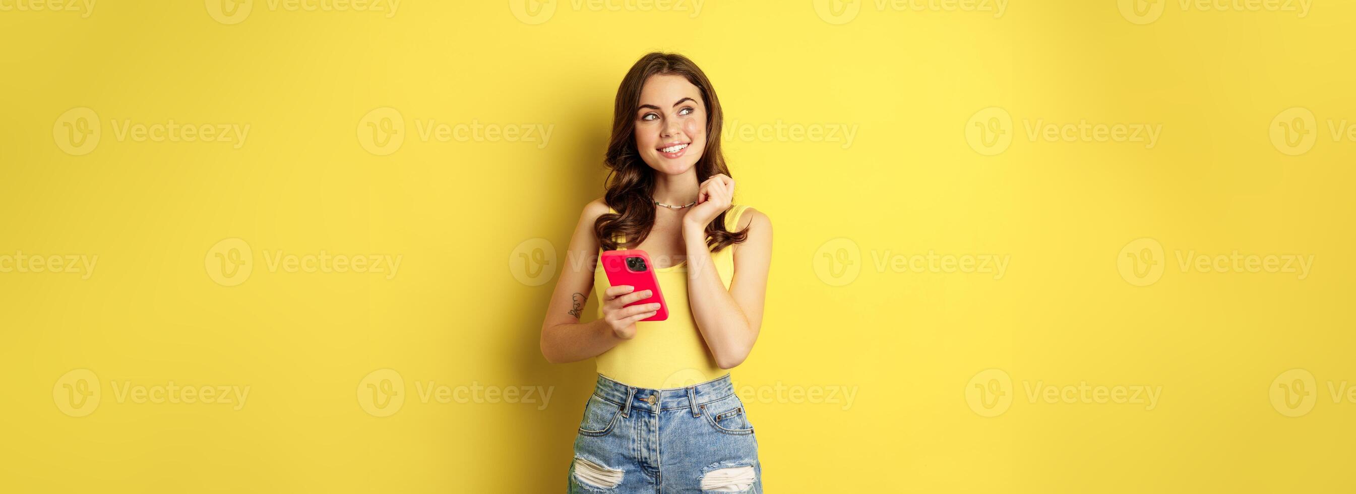 Technology and cellphone. Beautiful stylish girl order in online store, praying in application, using smartphone app and thinking, making decision, standing over yellow background photo