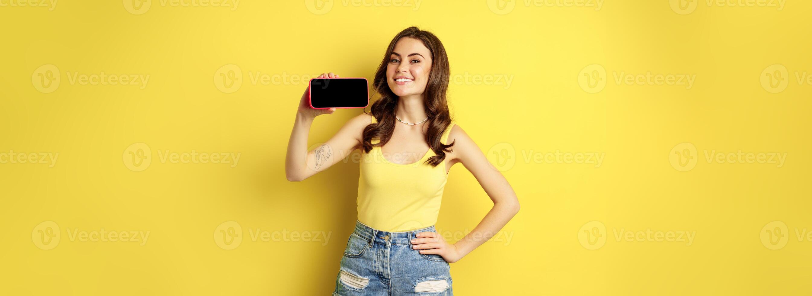 Proud smiling woman showing mobile phone horizontal screen, looking proud and satisfied, demonstrating smartphone display, app or online shopping store, yellow background photo