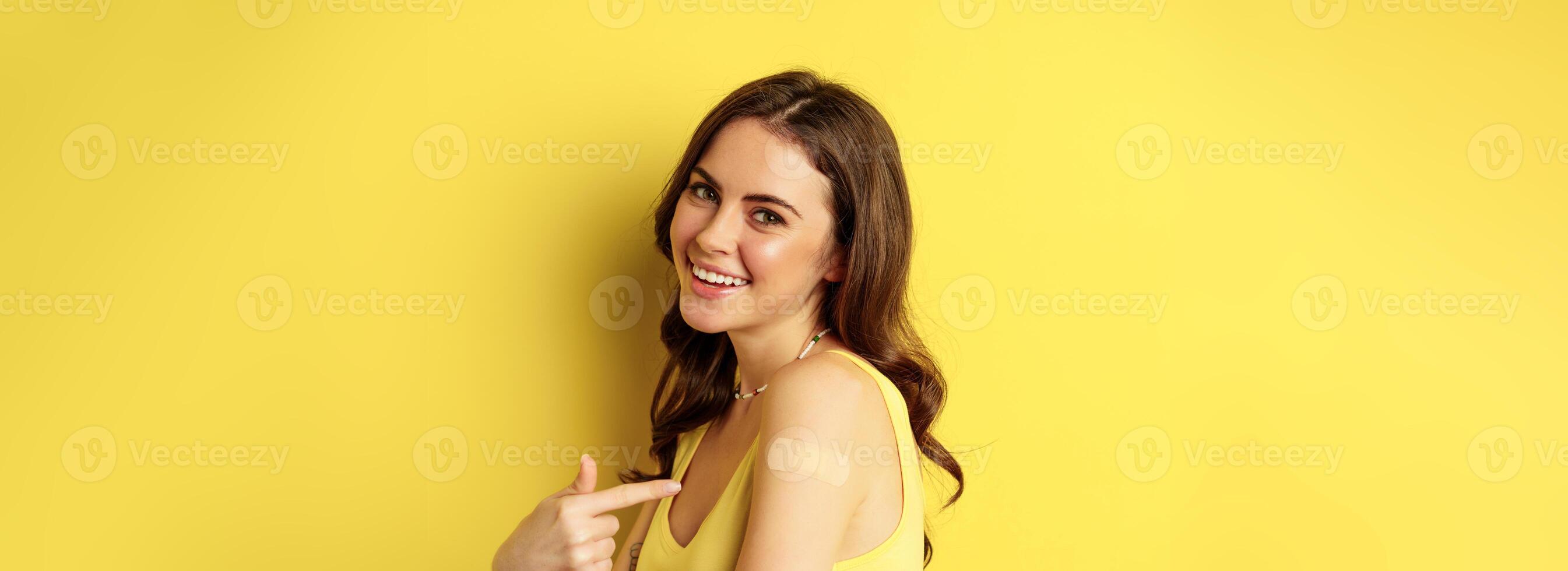 Close up of happy smiling girl with patch on shoulder after vaccination, covid-19 vaccinating campaign, standing over yellow background photo