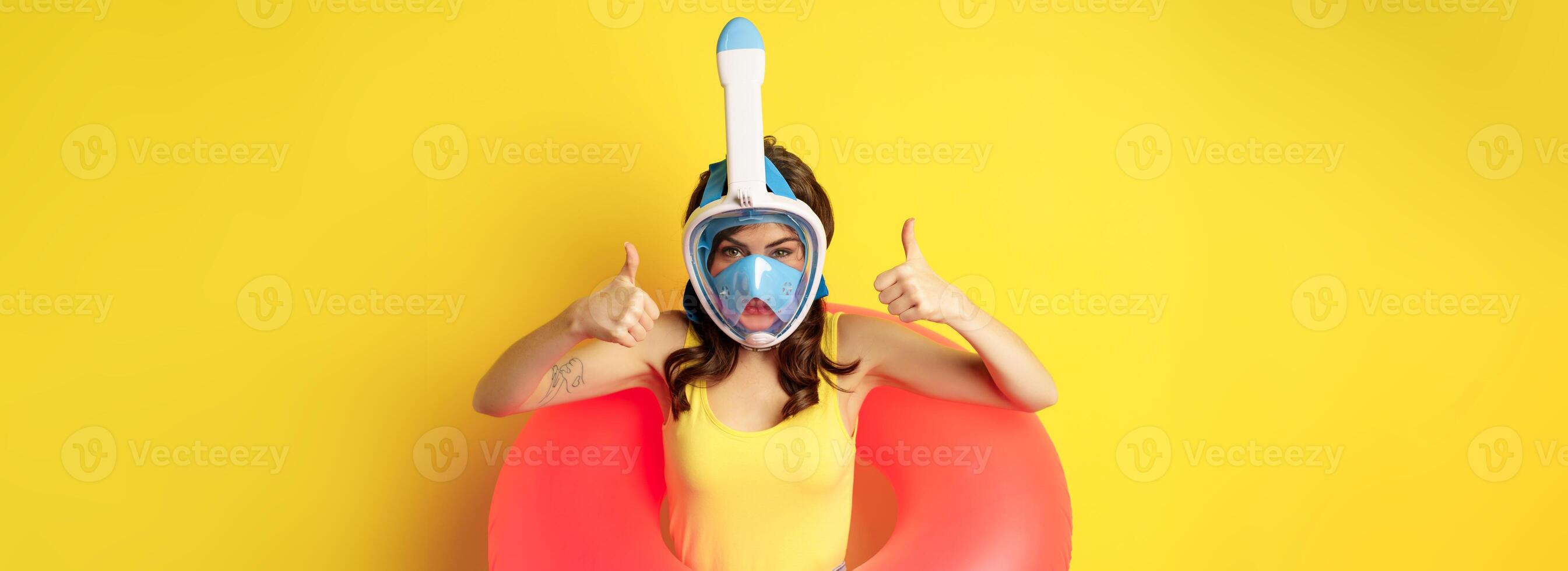 Funny happy woman in swimming ring, wearing snorkling mask for diving, showing thumbs up, good approval gesture, posing against yellow background photo