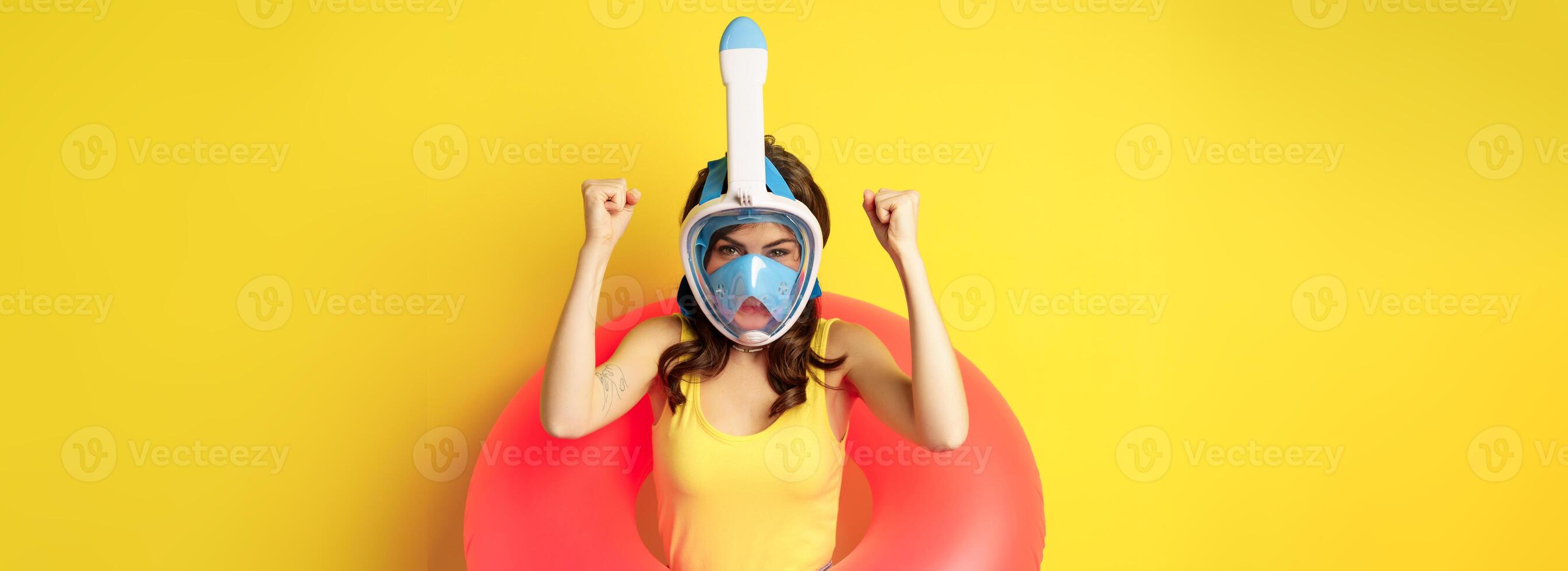 Portrait of enthusiastic young woman on vacation, wearing snorkling, diving mask and swimming ring, standing over yellow background photo