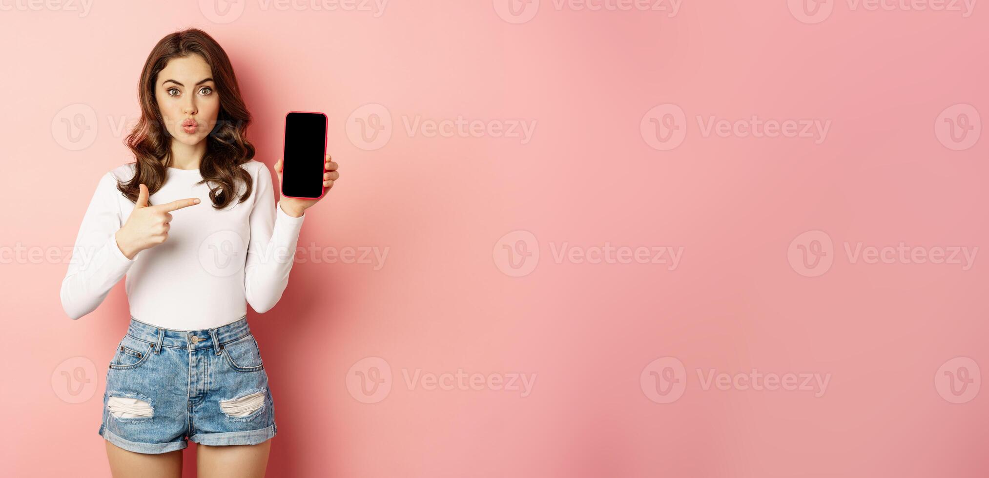 Surprised brunette girl pointing at mobile phone app screen, showing smartphone advertisement, online shopping offer, standing amazed against pink background photo