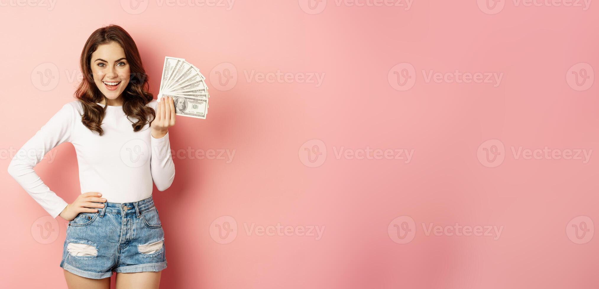 Attractive young woman holding money, cash in hands, concept of loans, microcredit and shopping, standing over pink background photo