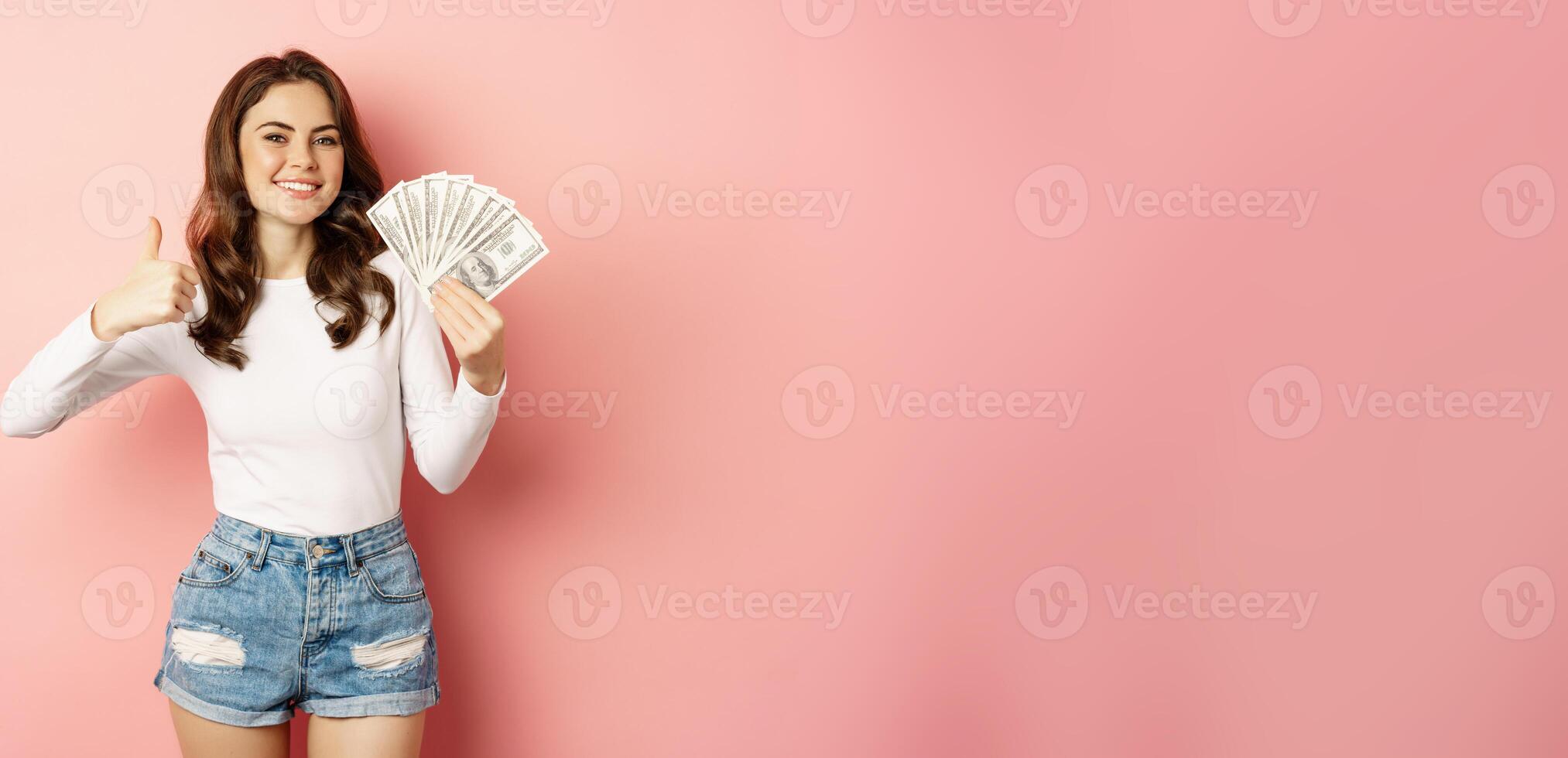 Easy money and loan concept. Smiling beautiful woman showing money with satisfied face, showing thumbs up, standing against pink background photo