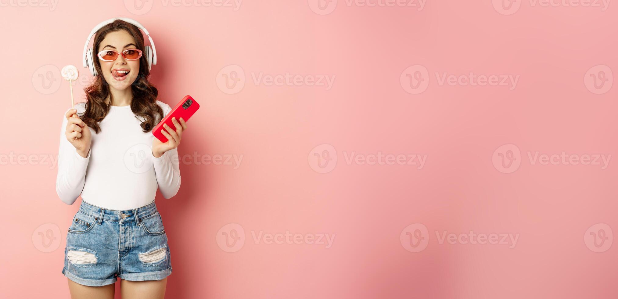 Stylish brunette woman dancing in headphones with mobile phone, listening music in earphones, licking lolipop, standing over pink background photo