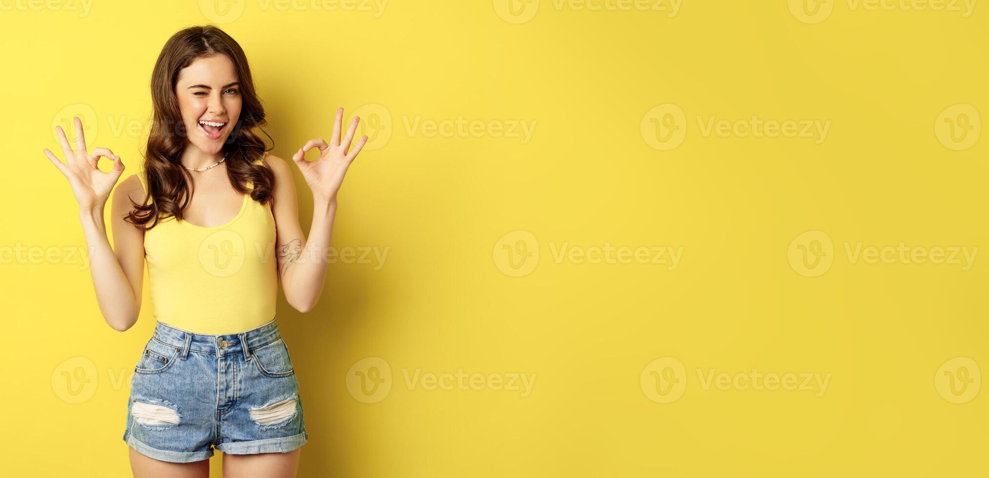 Smiling attractive woman showing okay signs and winking, assure you, praise and compliment smth good, recommending, standing over yellow background photo