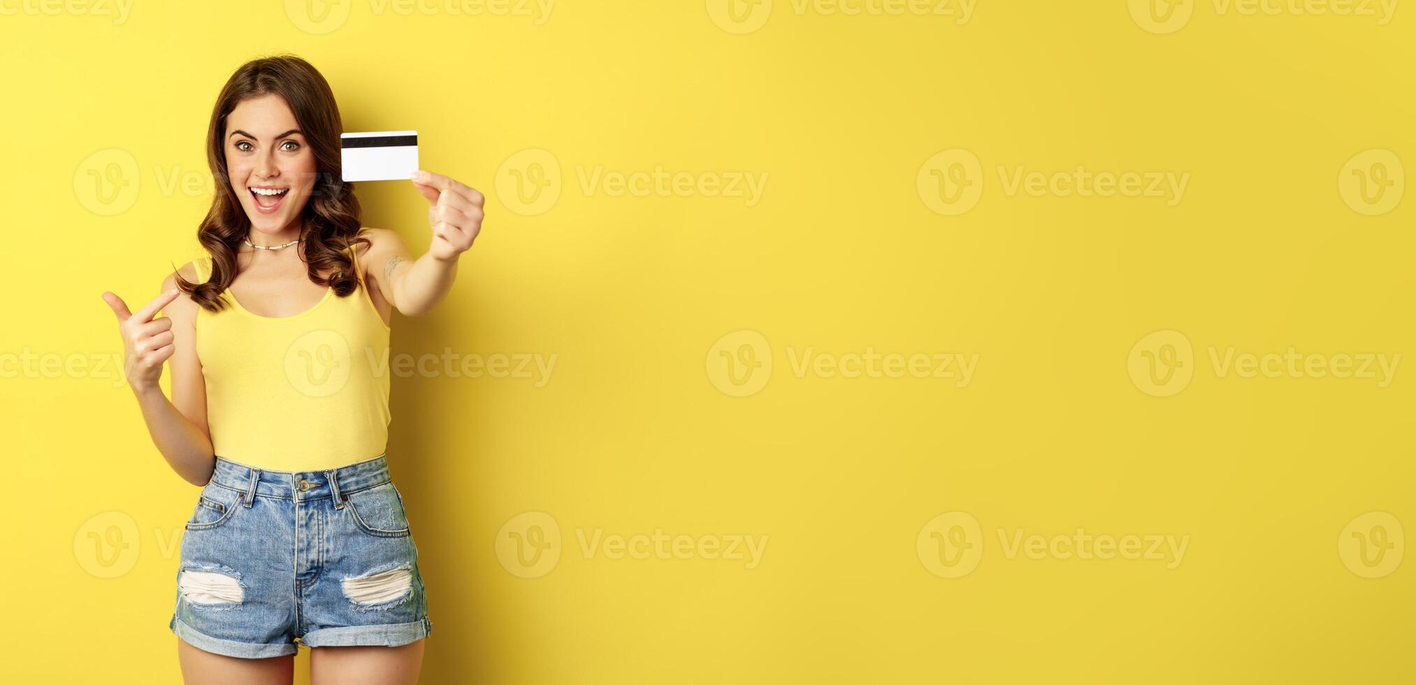 Portrait of brunette girl showing her credit card and smiling, wearing summer clothes, concept of bank, money and shopping, standing over yellow background photo