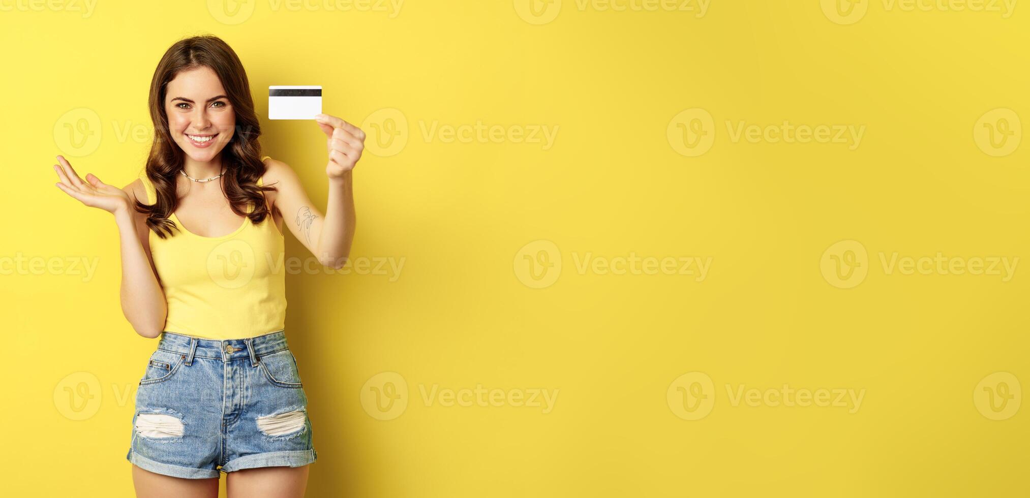 Portrait of brunette girl showing her credit card and smiling, wearing summer clothes, concept of bank, money and shopping, standing over yellow background photo