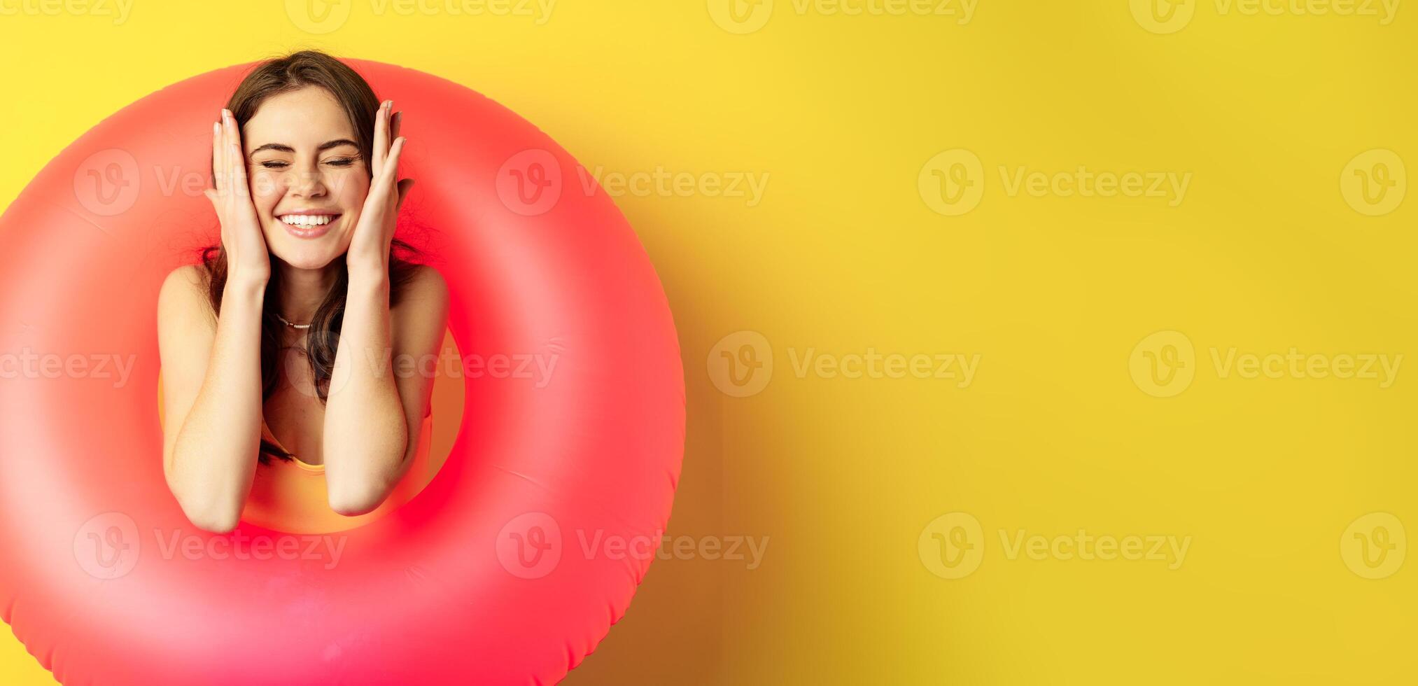 Happy beautiful young woman relaxing on summer vacation, wearing pink swim ring for beach holiday, standing against yellow background photo