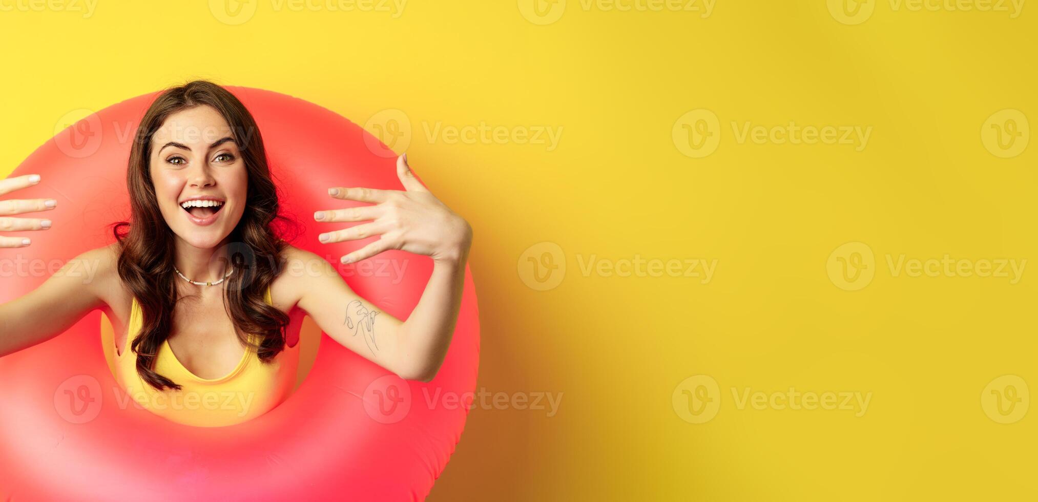 Close up portrait of beautiful happy woman inside pink swimming ring, luring, inviting people on summer vacation, beach party, standing over yellow background photo
