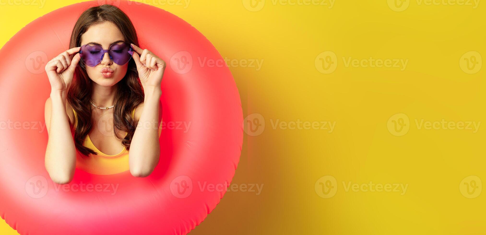 Close up portrait of stylish young woman, girl with swimming ring and summer sunglasses, ready for beach holiday, vacation, standing over yellow background photo