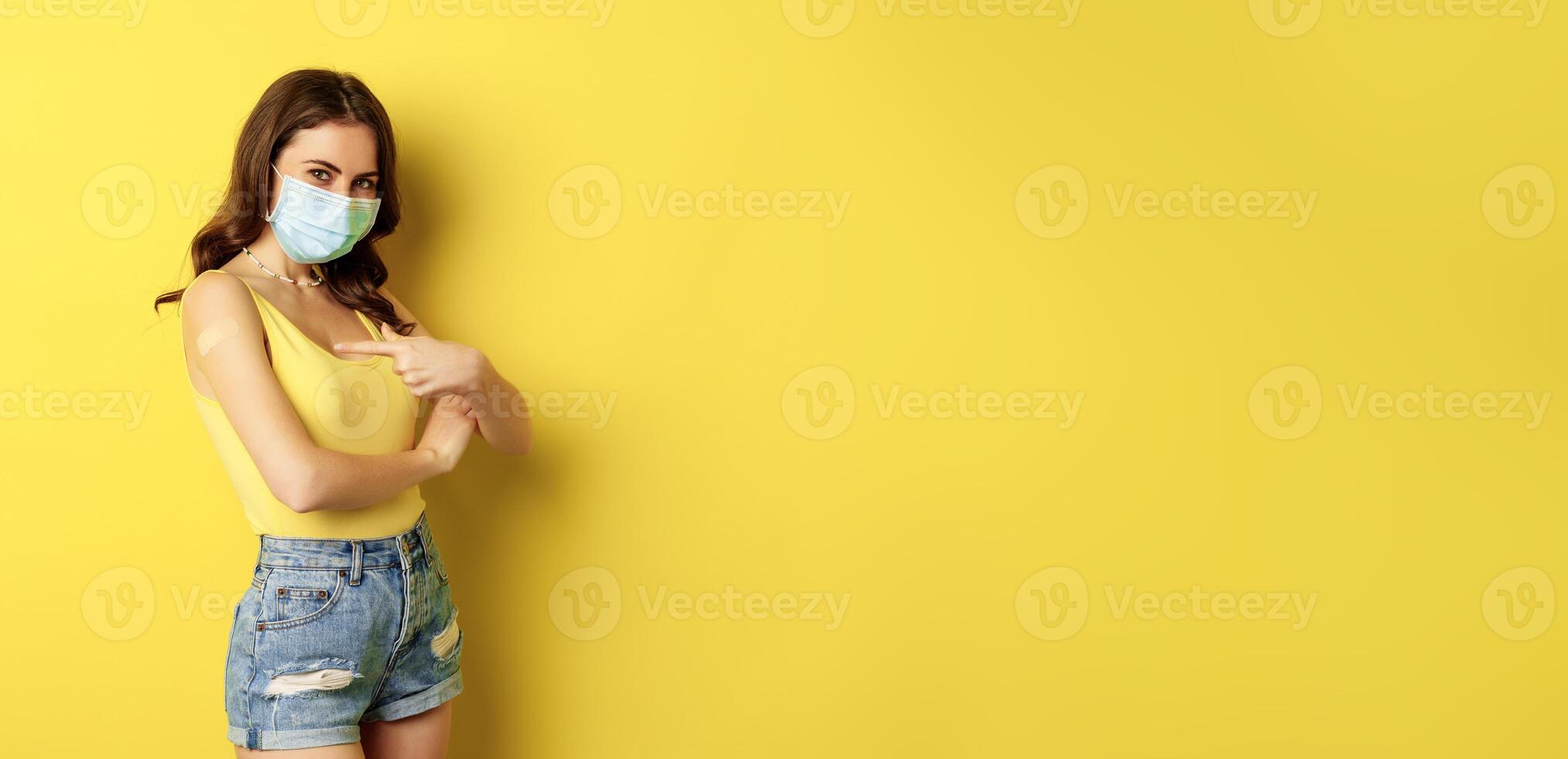 Covid-19 vaccination. Beautiful young woman in face medical mask got vaccinated during pandemic, standing over yellow background photo