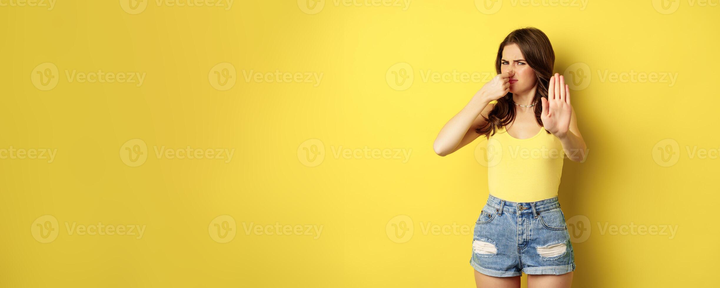 Woman showing stop sign and shut nose from disgust, dislike bad smell, declining something awful, standing over yellow background photo